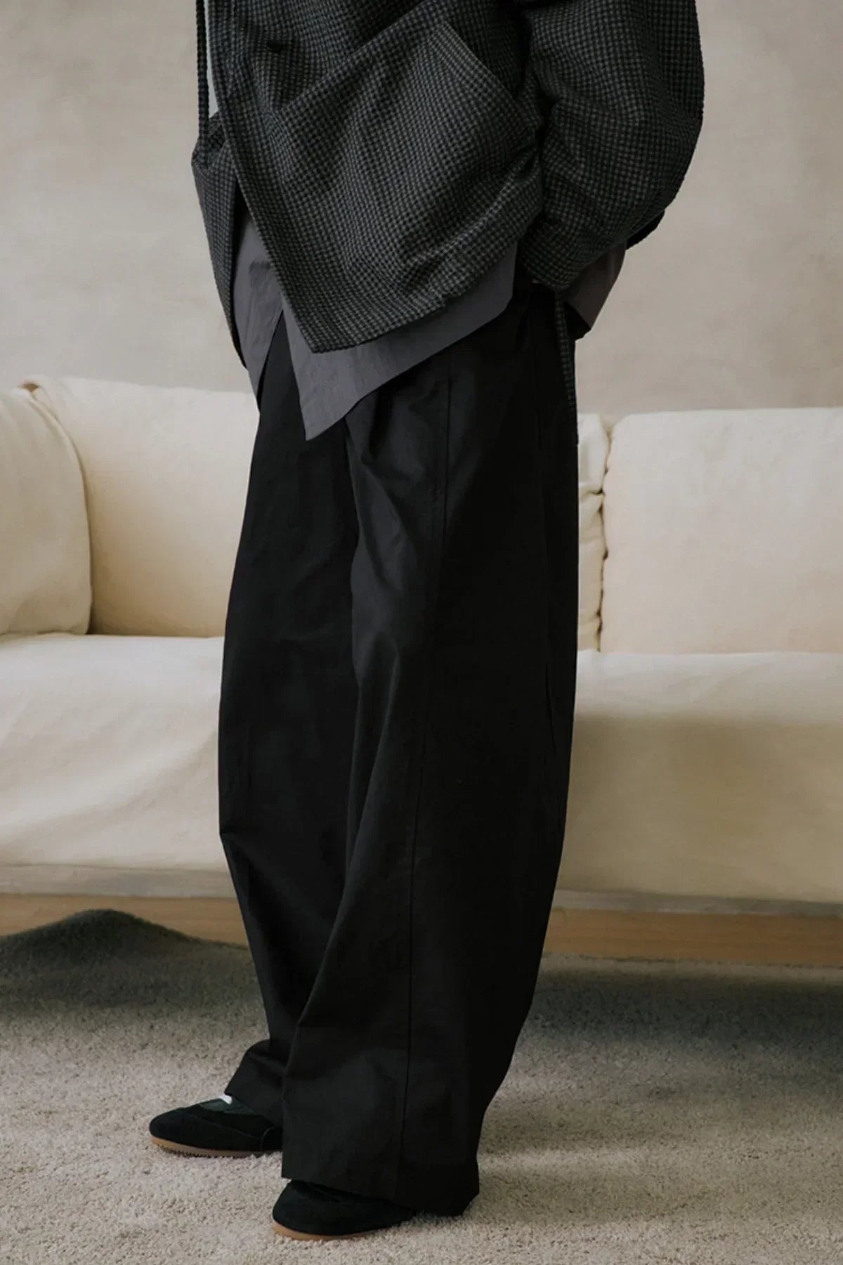 Person wearing black unisex wide-leg pants with layered tops standing on carpet in front of beige sofa