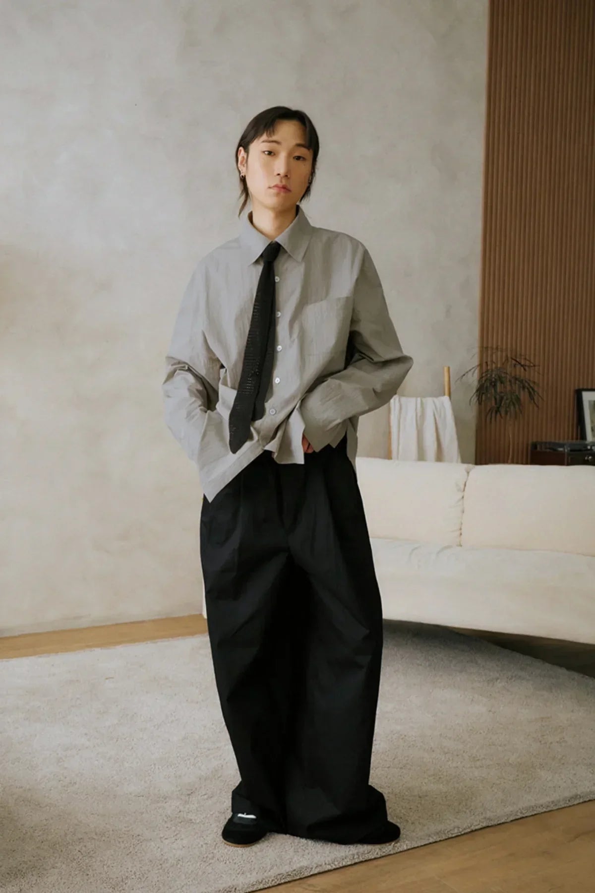 Person wearing loose black pants, gray shirt, and black tie standing in a modern living room