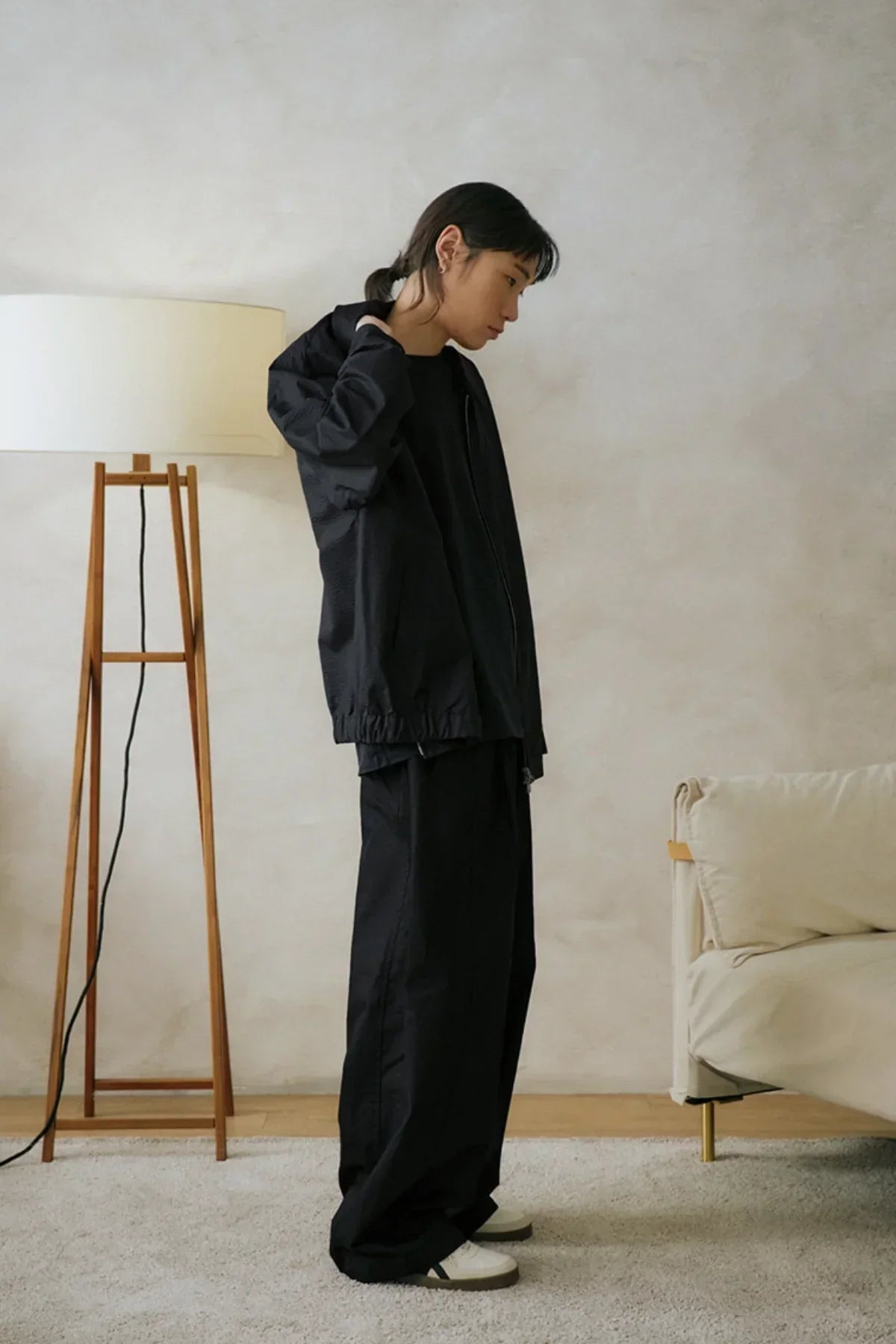 Side view of a model wearing black unisex loose pants and matching jacket indoors on carpeted floor next to a wooden lamp and beige sofa