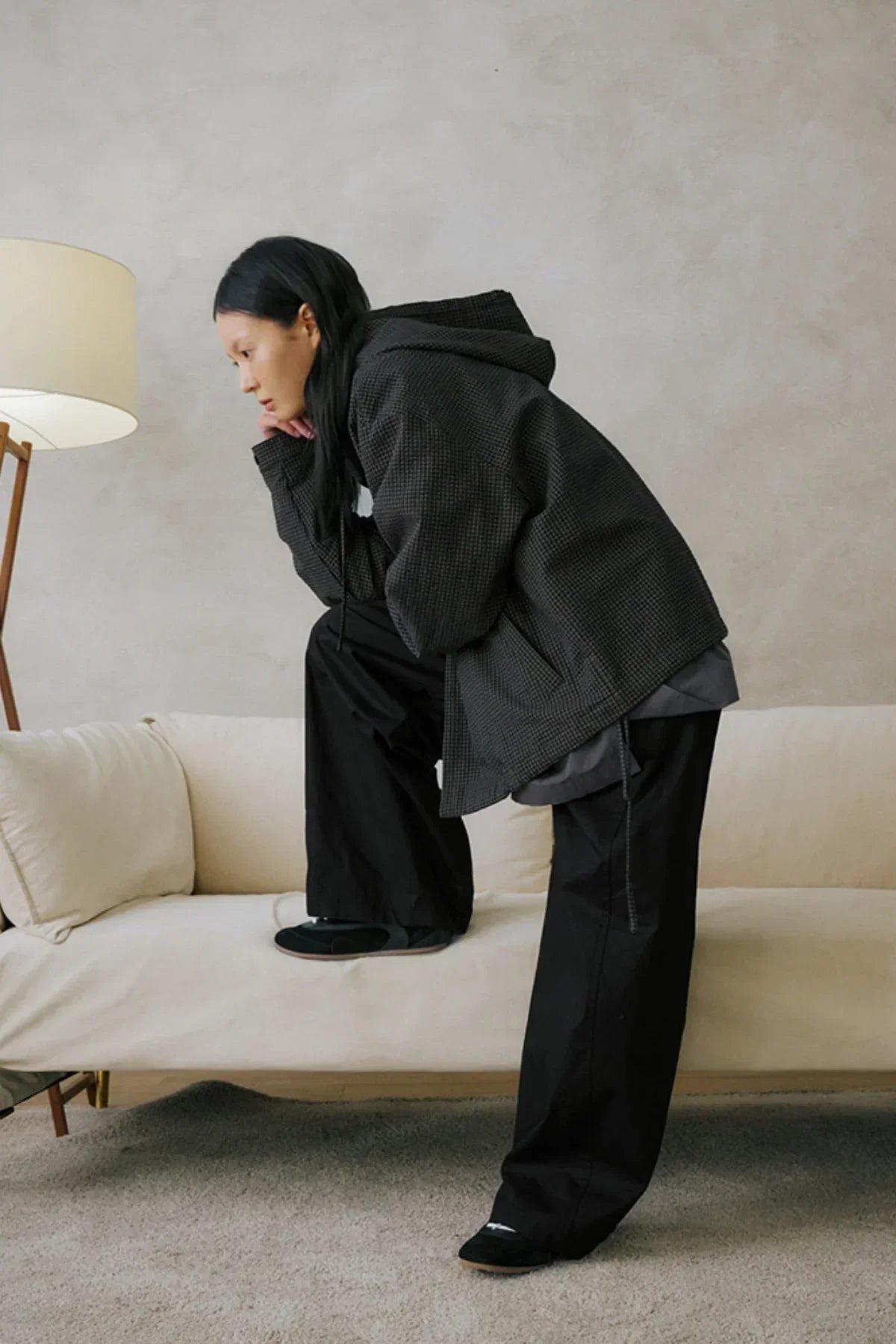 Person wearing black wide-leg pants and checkered hoodie posed on beige sofa in minimalist room