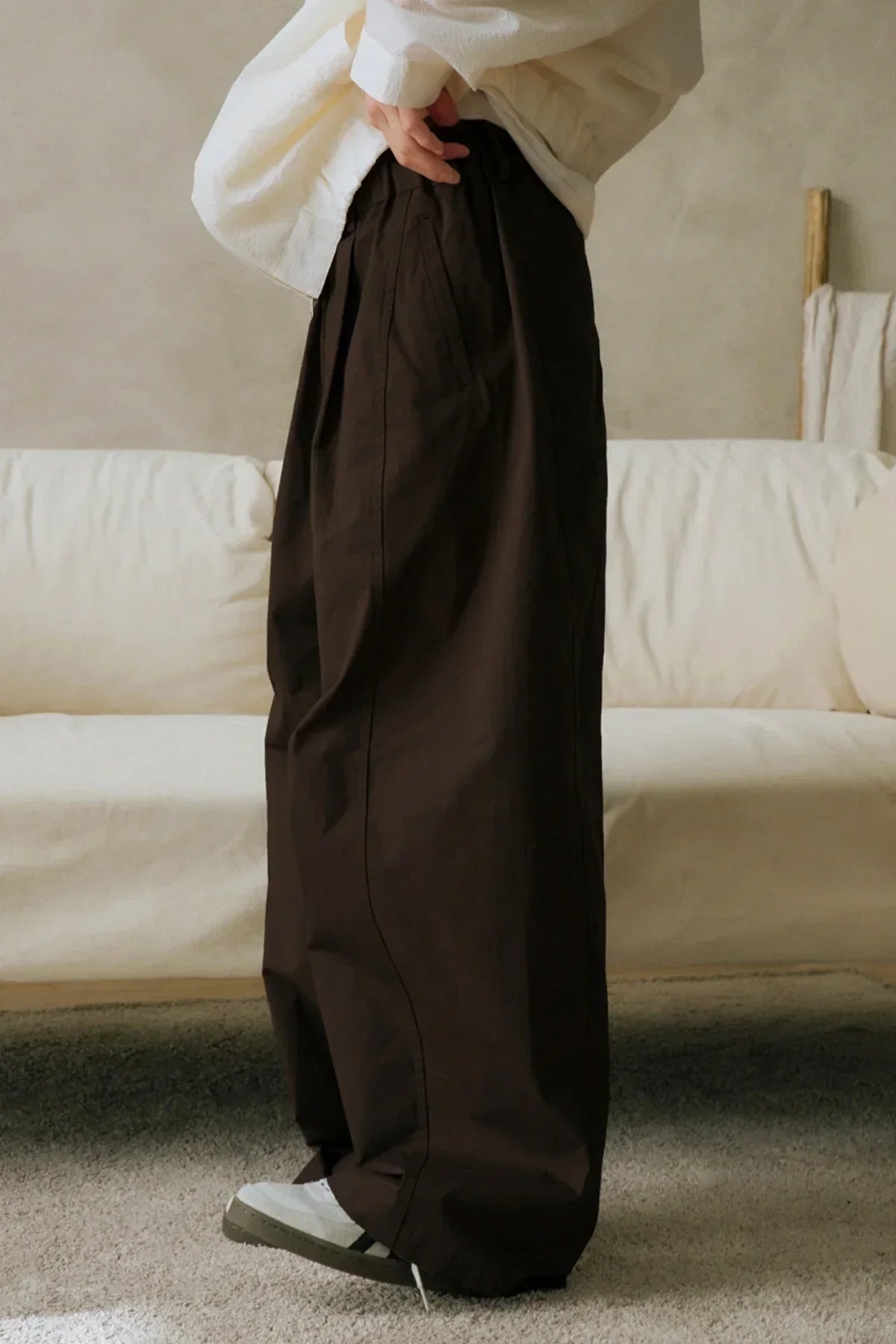 Side view of a person wearing wide-leg brown unisex pants and white sneakers in a cozy living room