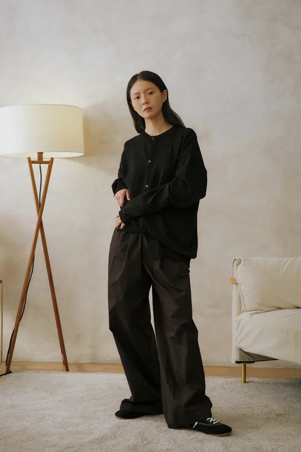 Woman wearing loose dark brown unisex pants and black button-up top standing in minimalist living room