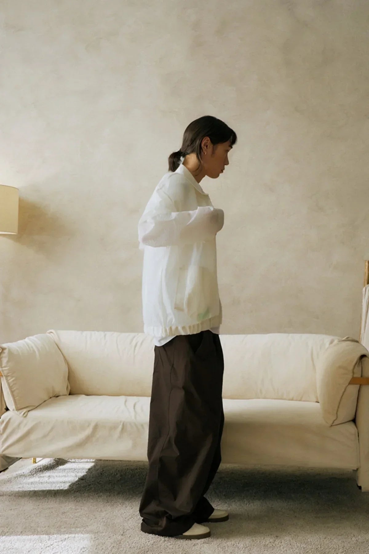 Model in white jacket and loose brown unisex pants standing indoors by cream sofa and beige wall