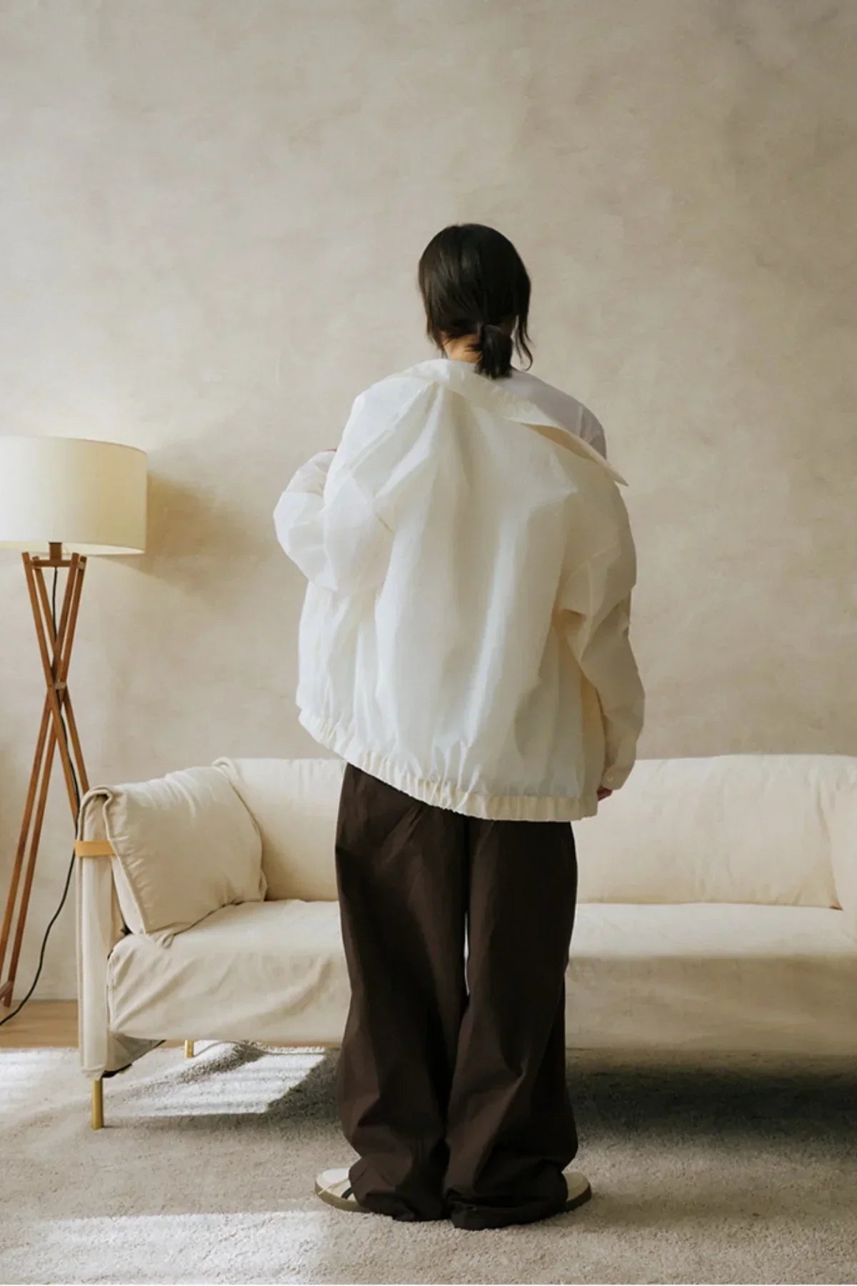 Person wearing white jacket and loose brown pants standing in minimalist living room with beige sofa and wooden floor lamp