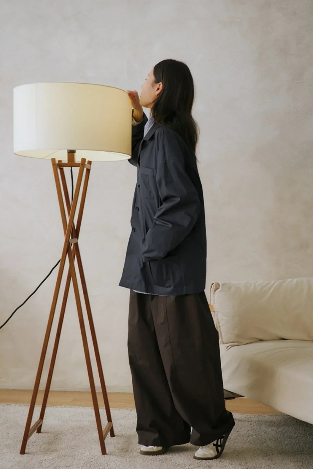Person wearing dark oversized jacket and wide brown pants standing near wooden tripod floor lamp in minimal room