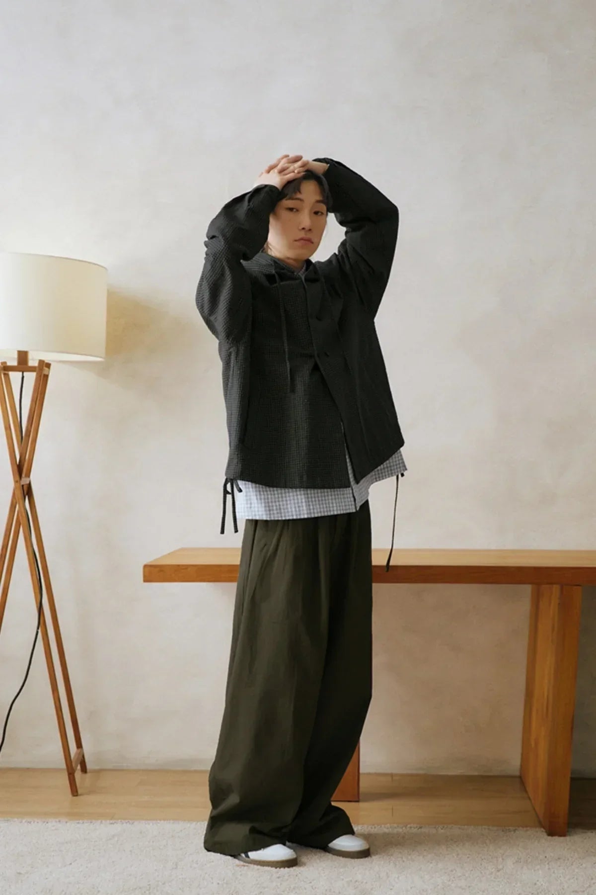 Man wearing black jacket and loose khaki pants standing indoors next to wooden bench and floor lamp