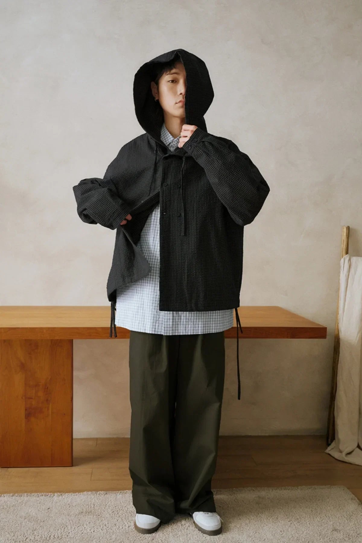 Model wearing black hooded jacket, checked shirt, loose khaki pants, and white sneakers in minimalist room