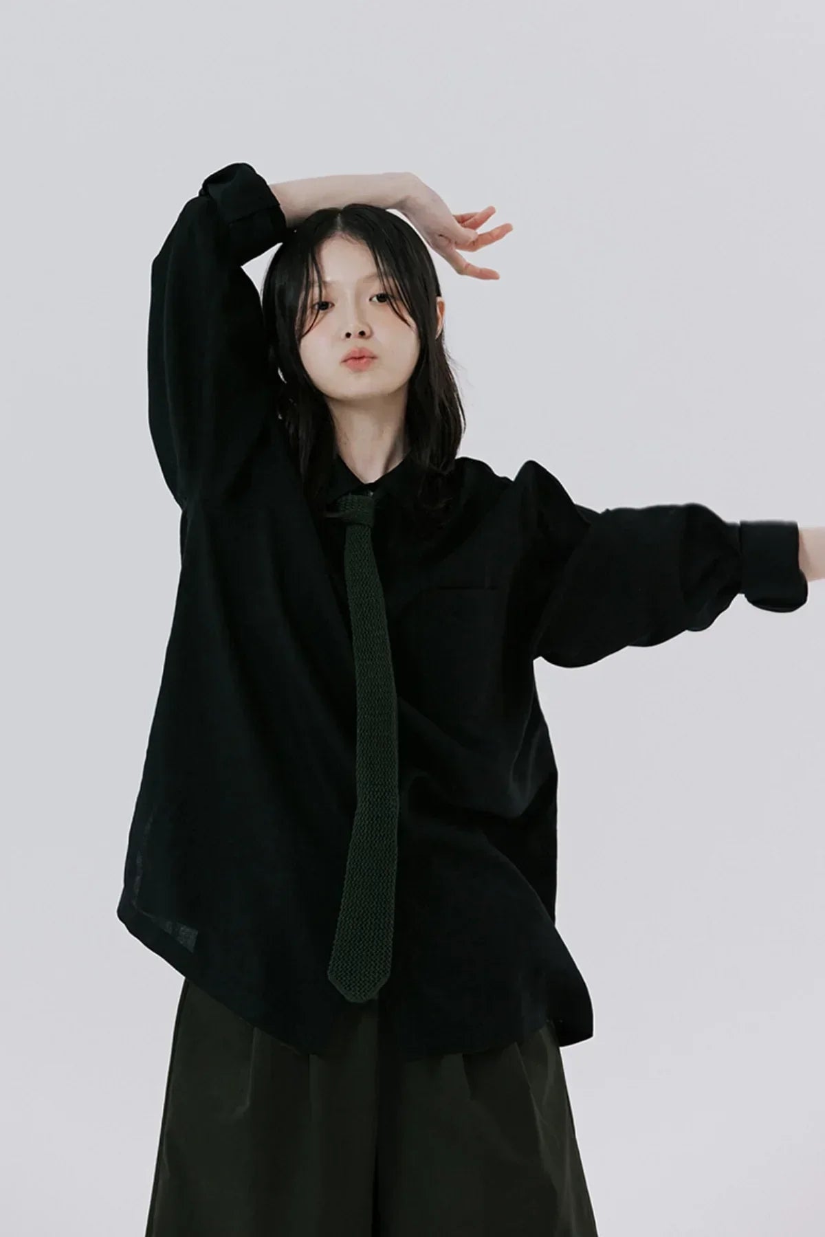 Young woman posing in loose black shirt and dark tie against plain gray background