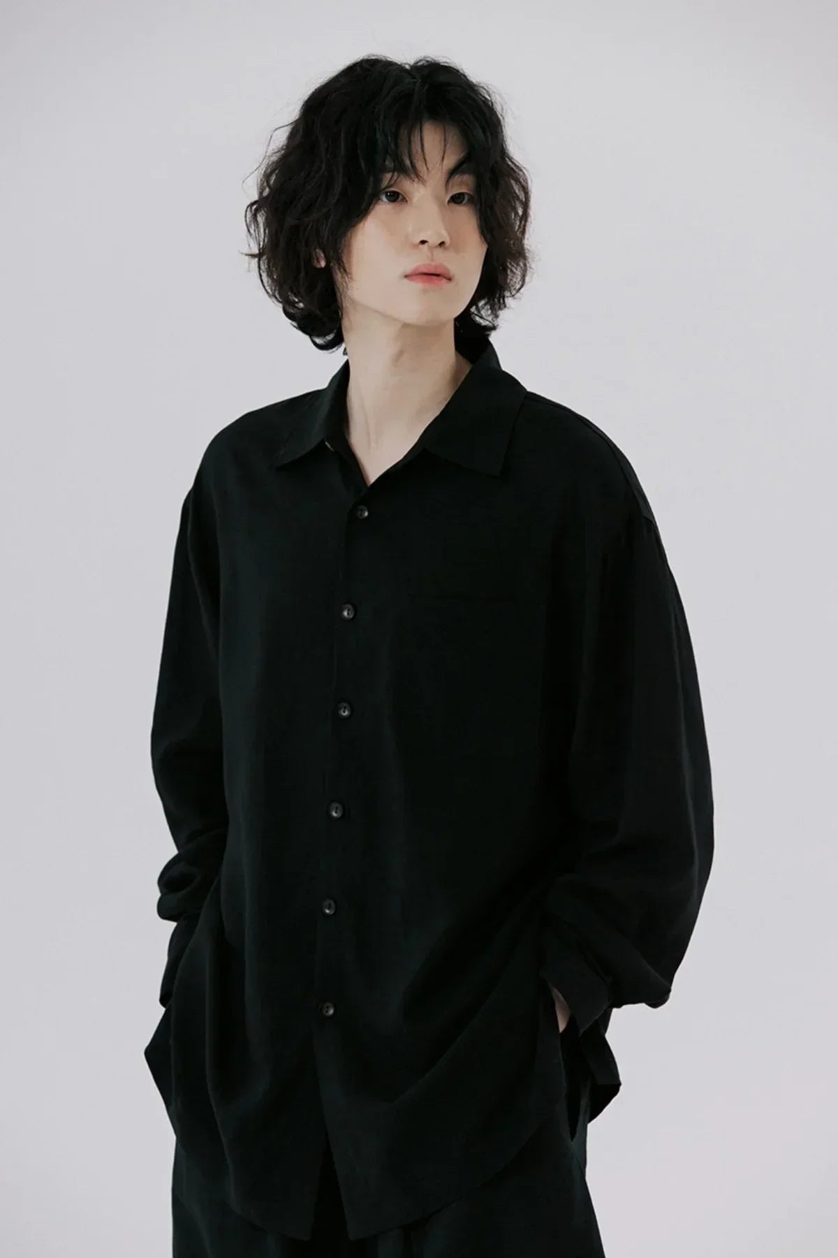 Young person with curly hair wearing an oversized black button-up shirt with hands in pockets