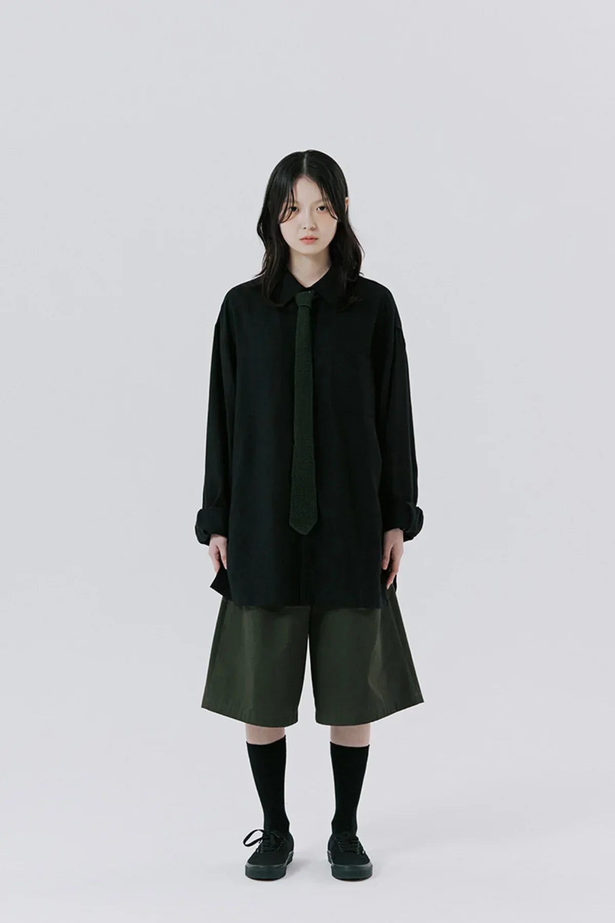 Young woman wearing oversized black shirt, dark green tie, and wide khaki shorts on plain background