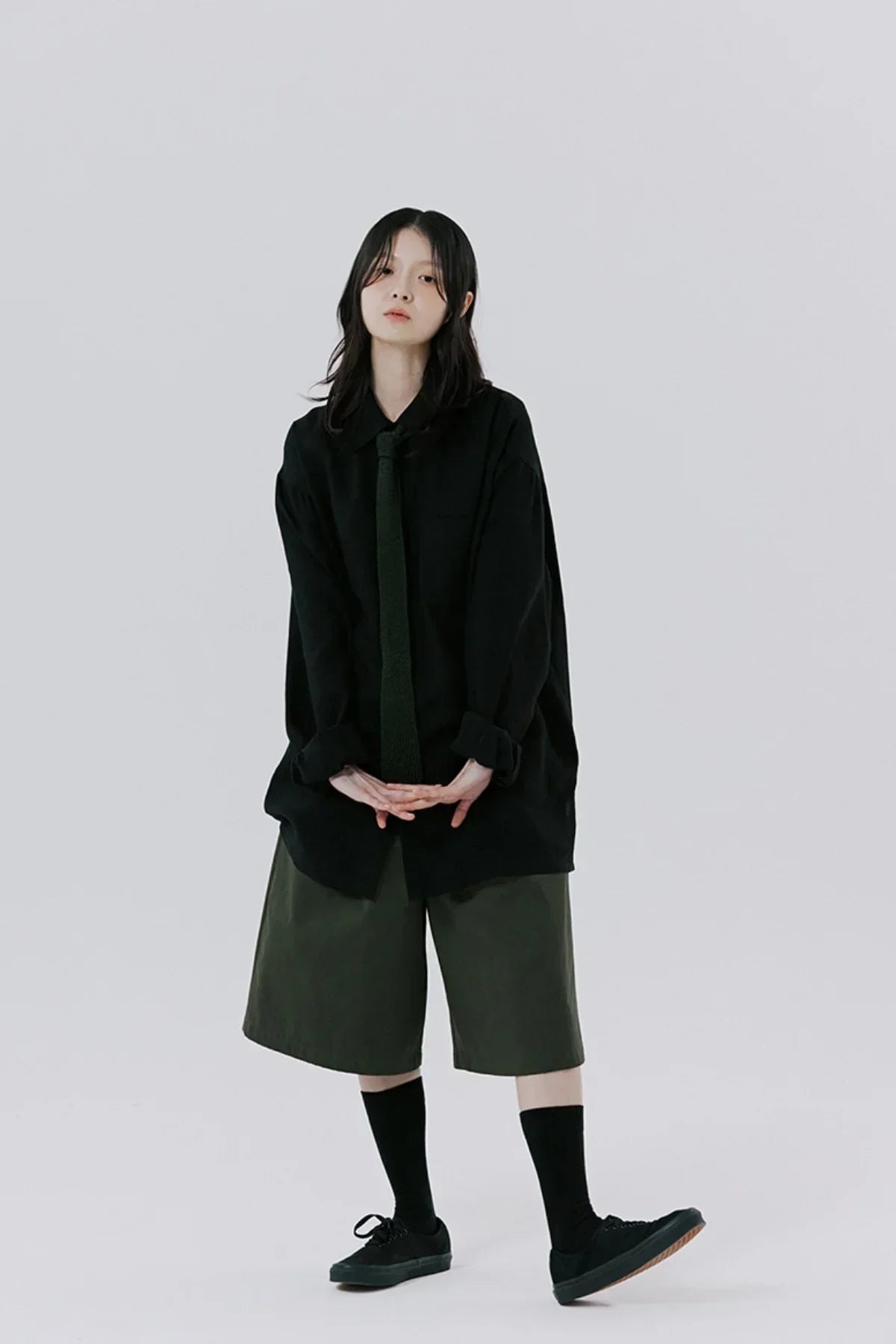 Young woman in oversized black shirt with black tie, olive green wide shorts, black socks, and black sneakers
