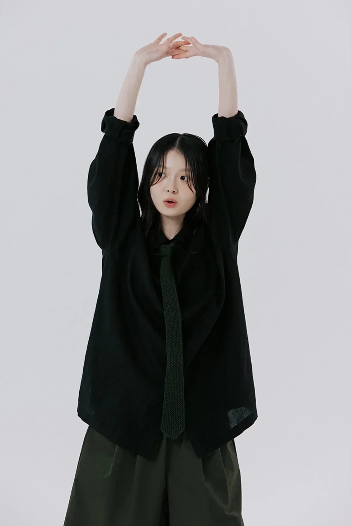 Young woman stretching in black oversized shirt and matching knitted tie with dark pants on plain background