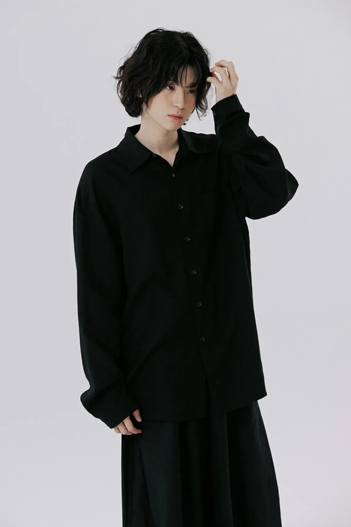 Young model with wavy hair wearing a loose black button-up shirt and black pants on gray background