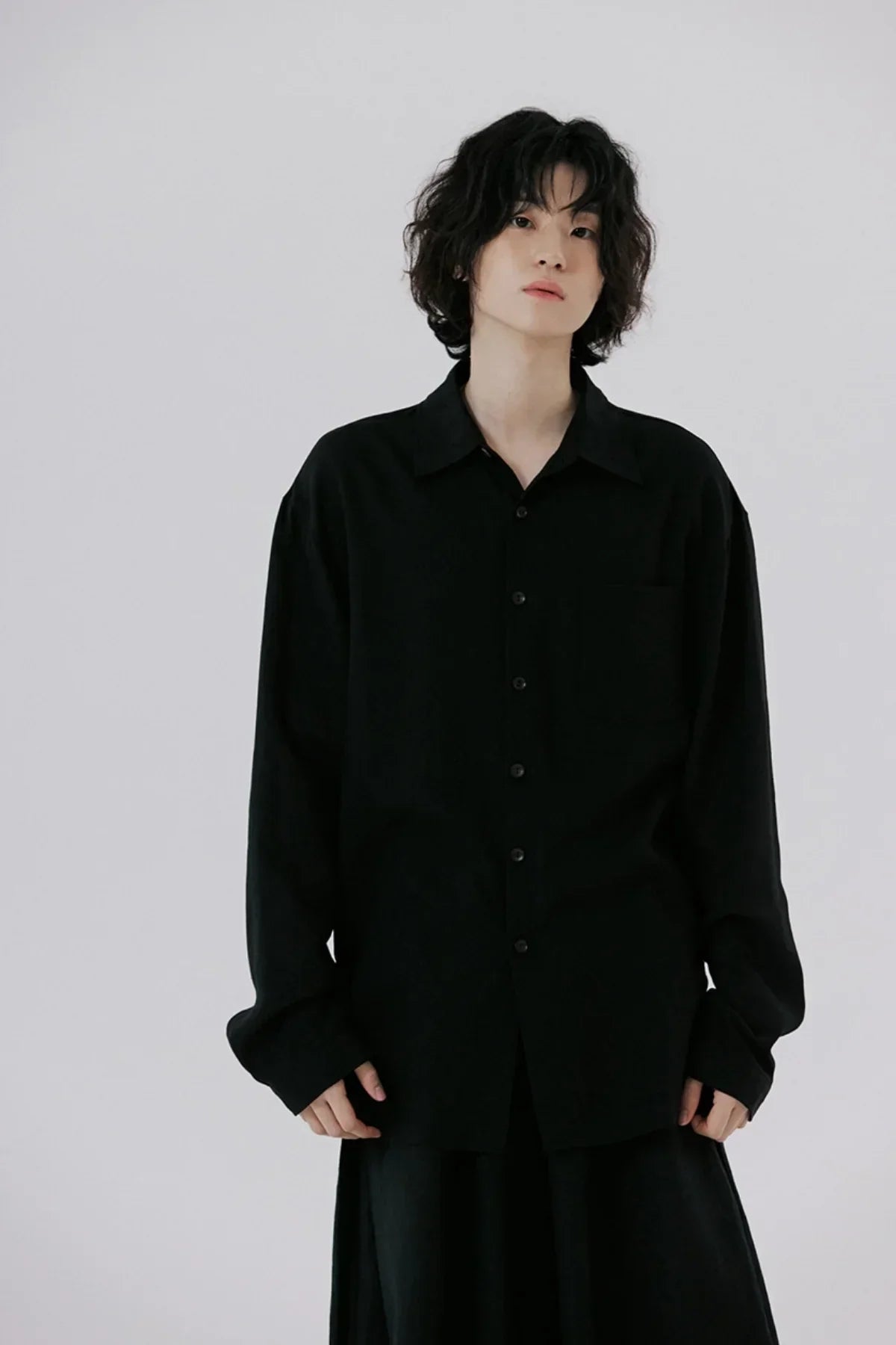 young person with curly hair wearing oversized black button-up shirt against plain gray background