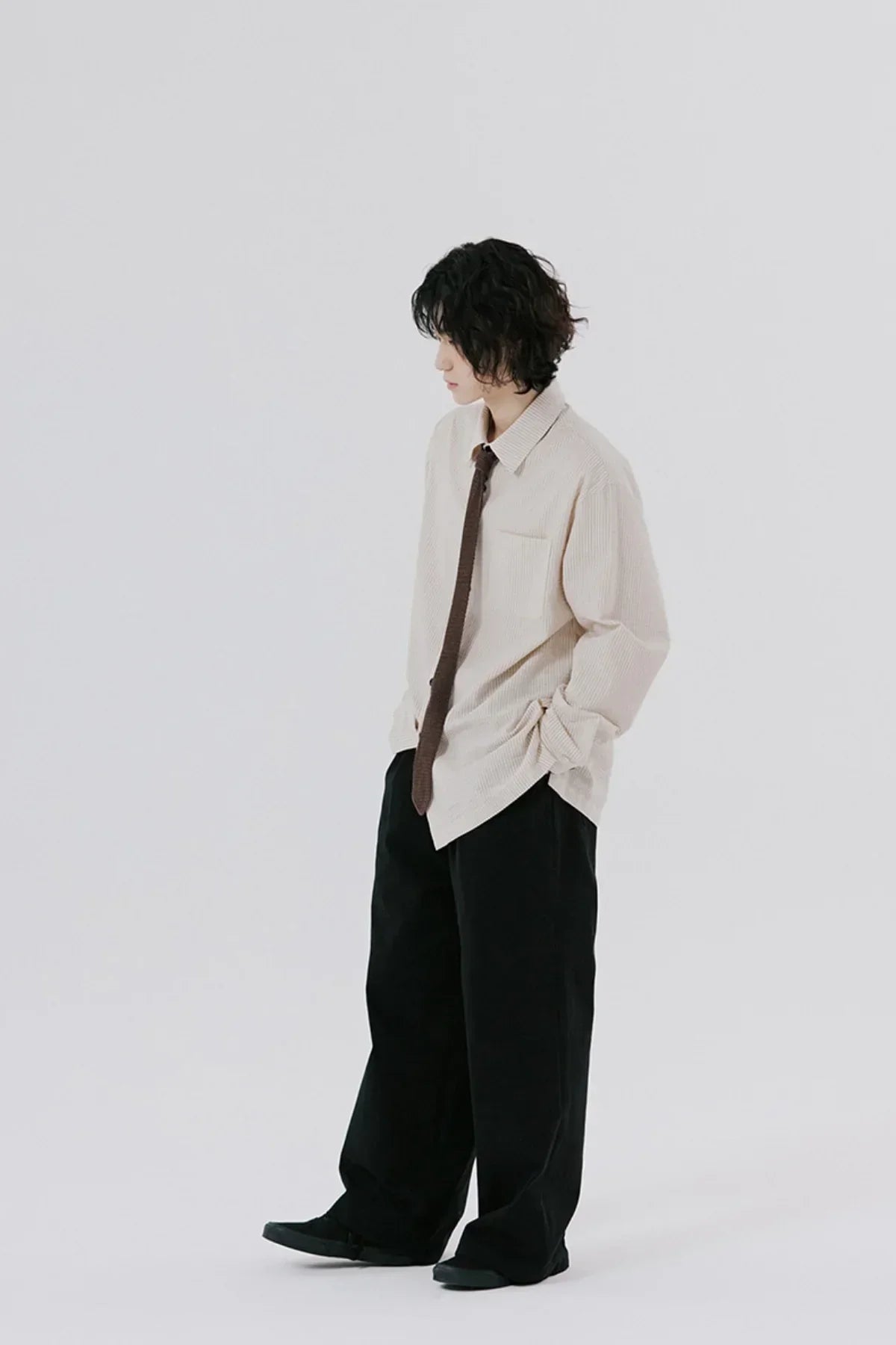 young person wearing beige long-sleeve shirt with tie and loose black pants on white background