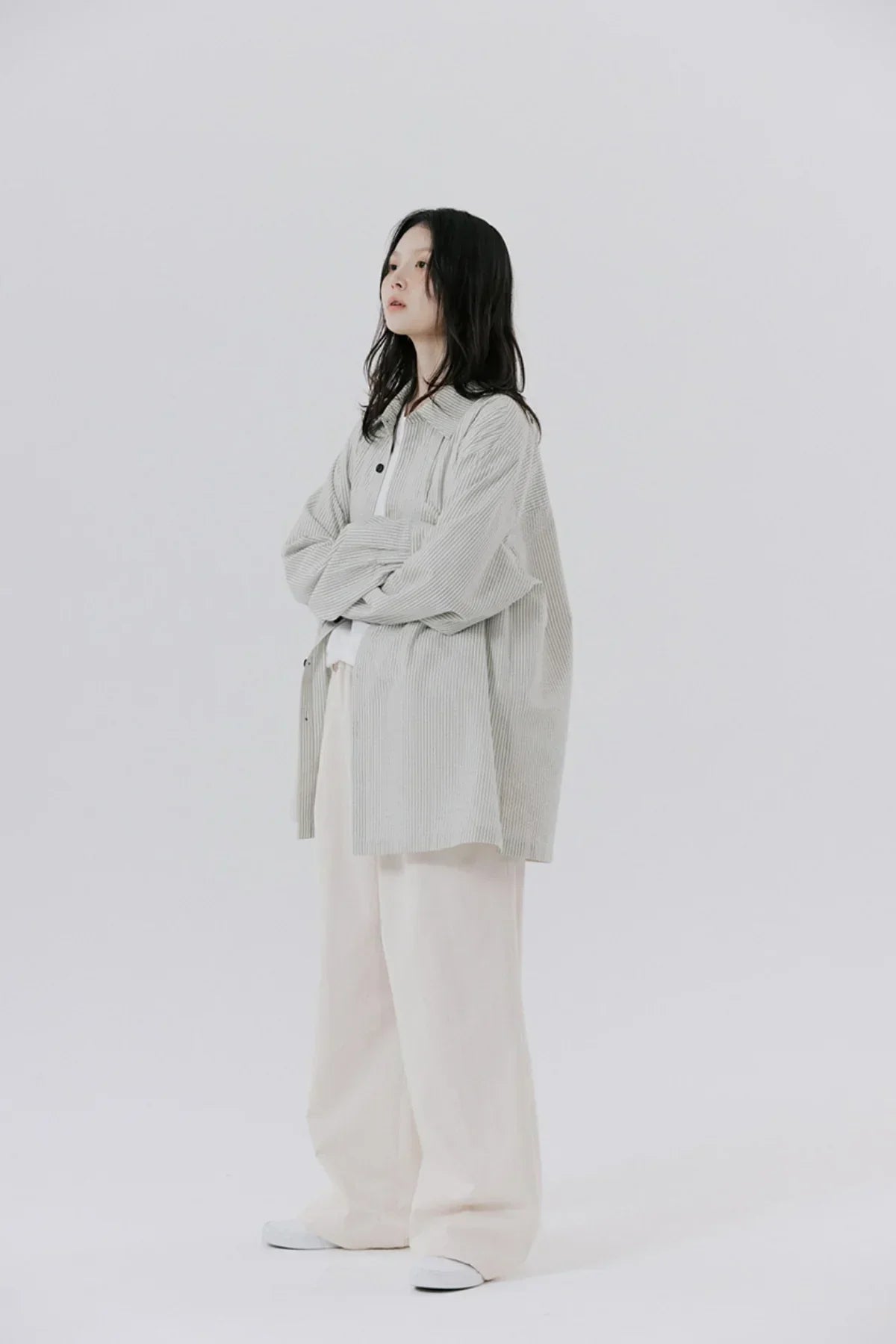 Woman wearing oversized light gray corduroy shirt, wide white pants, and white sneakers on plain background