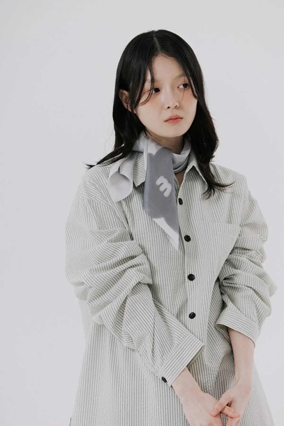 Young woman wearing oversized light grey striped shirt and grey scarf against white background