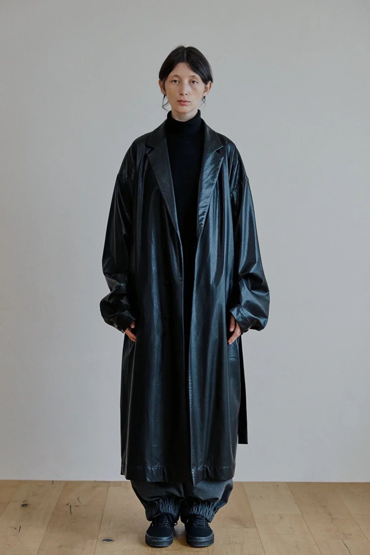 Model wearing a long black unisex leather trench coat with oversized sleeves, black turtleneck, dark pants, and black sneakers on wooden floor
