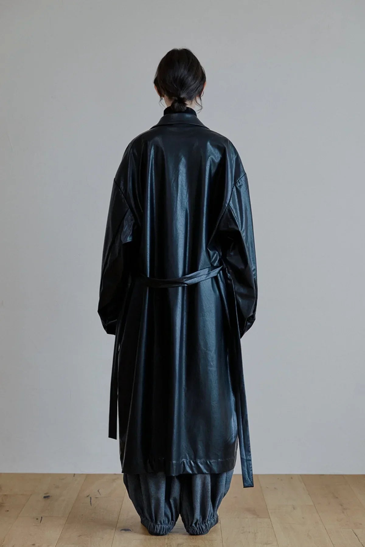 Back view of model wearing black unisex leather trench coat with belt and loose gray pants on wooden floor