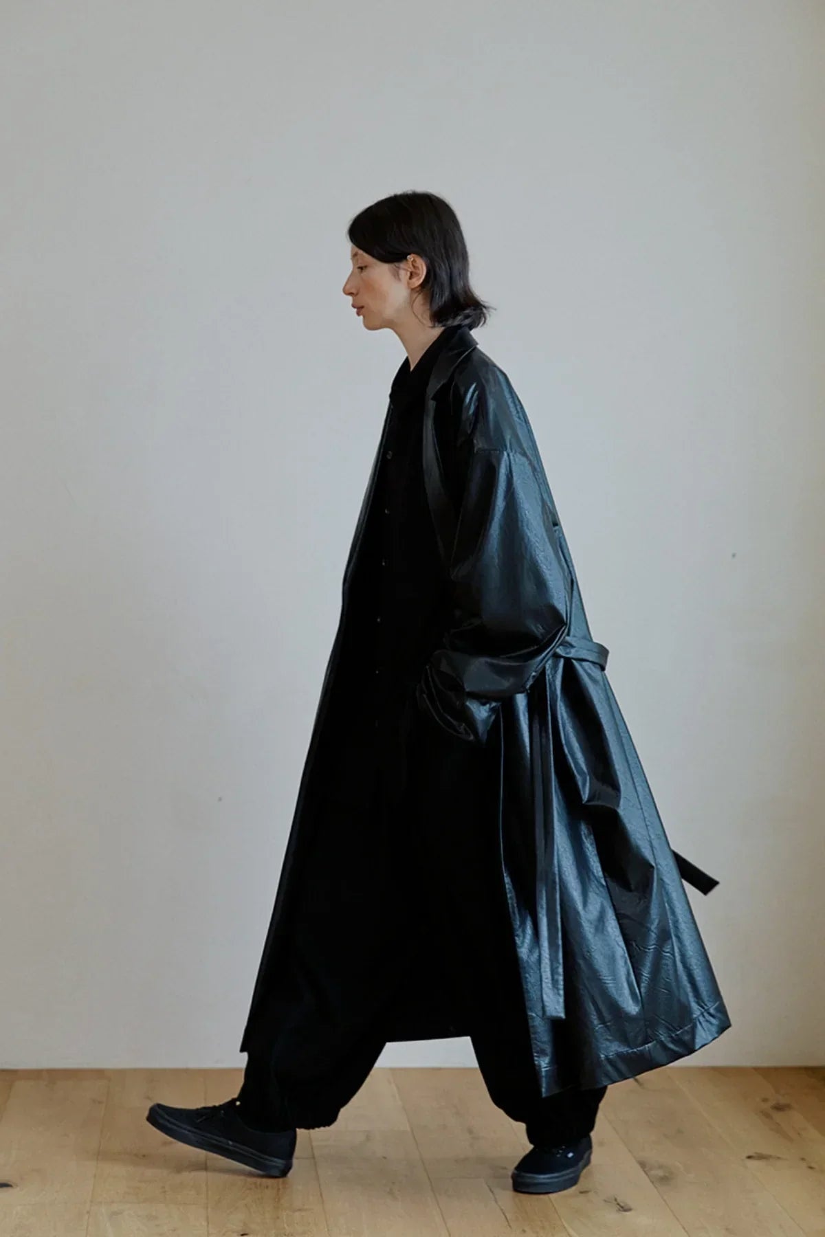 Model wearing a long black unisex leather trench coat and black sneakers walking indoors