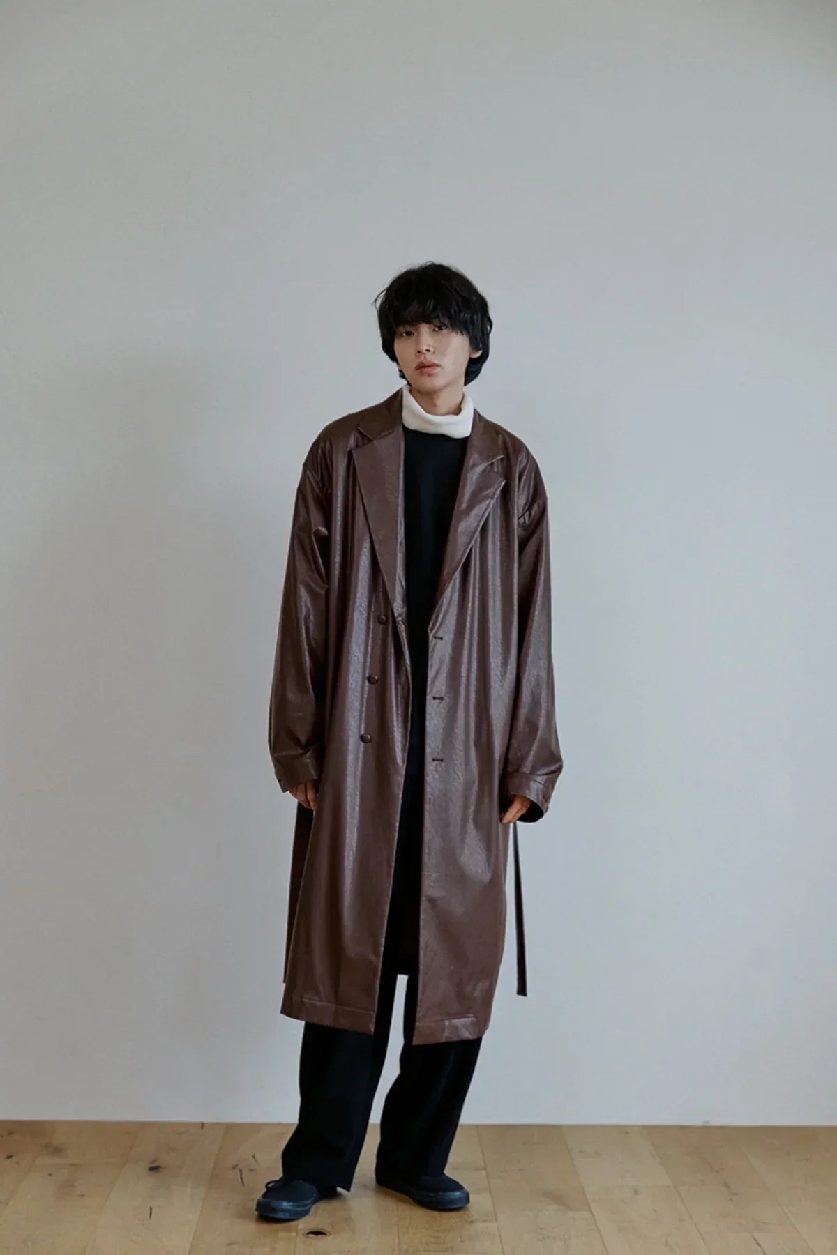 Model wearing unisex oversized brown leather trench coat on wooden floor against plain gray wall