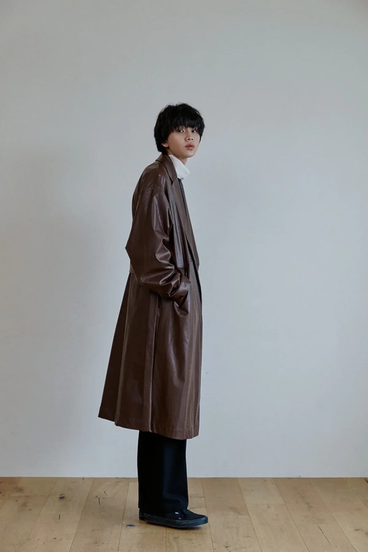 Model wearing unisex brown leather trench coat with black pants and black shoes on wooden floor