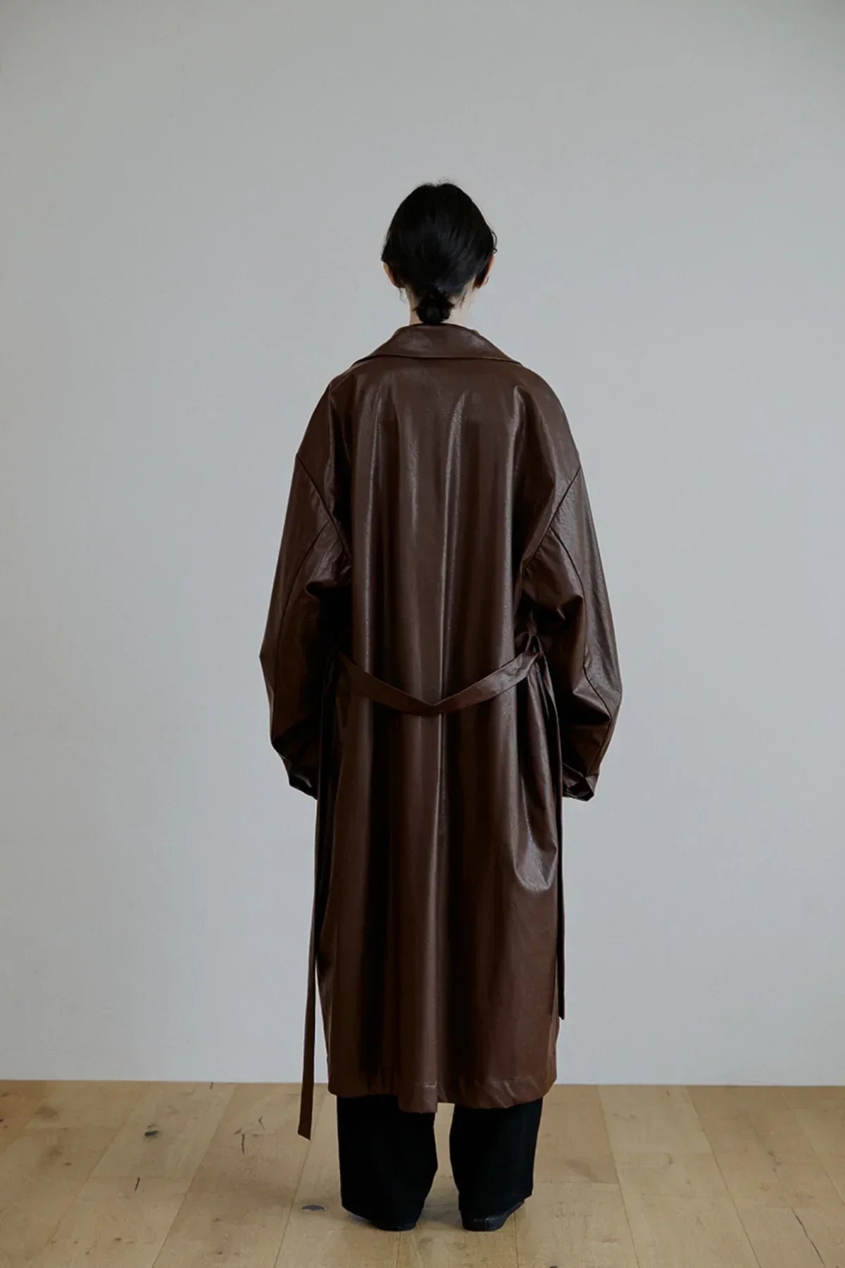 Back view of person wearing a long brown unisex leather trench coat indoors on wooden floor