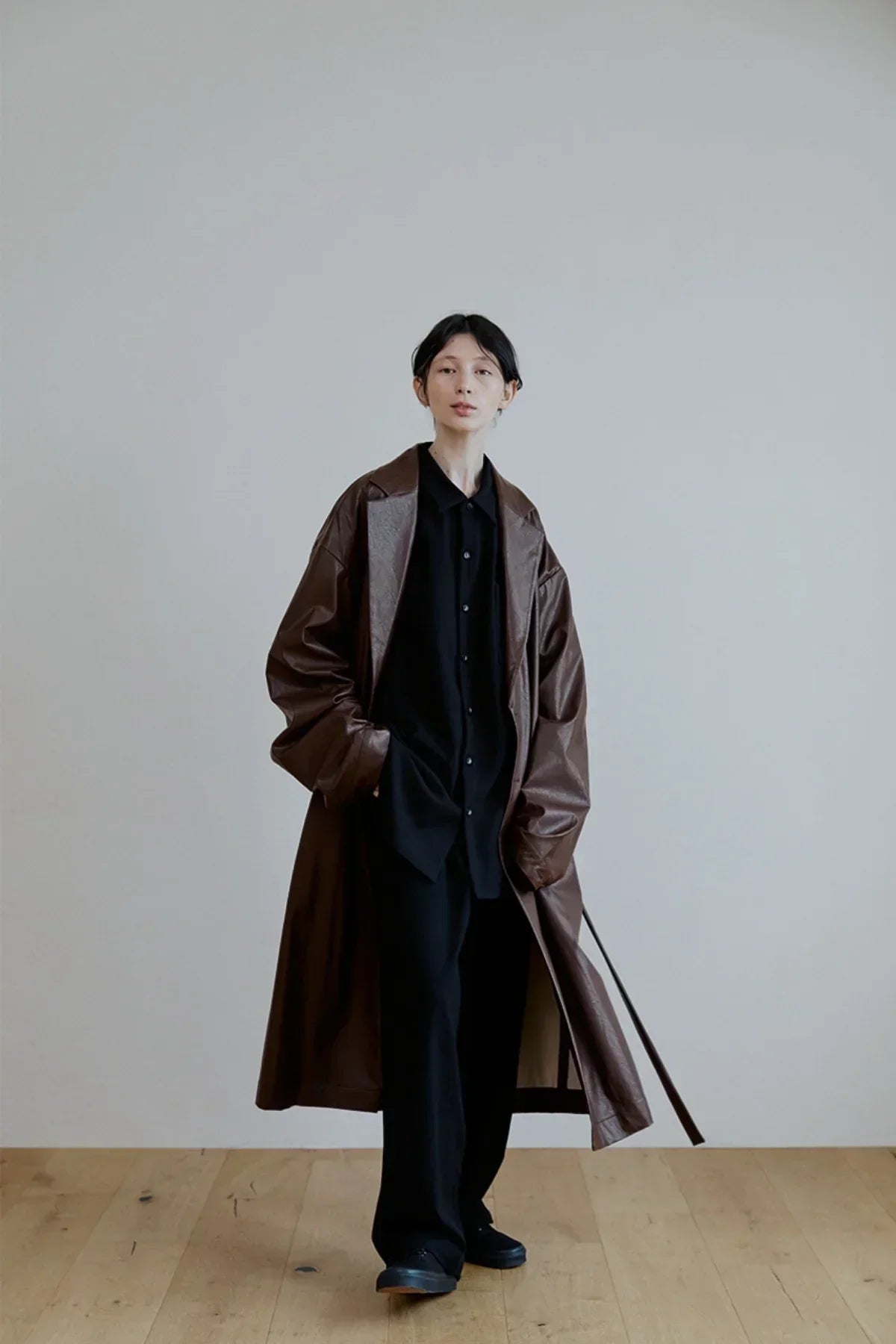 Model wearing oversized brown leather trench coat with black outfit on wooden floor and gray wall background