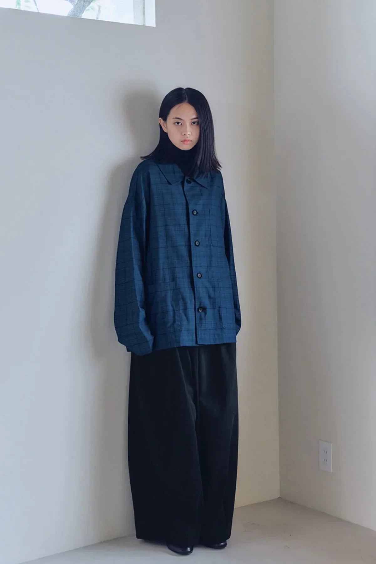 Model wearing wide black balloon pants and oversized blue checkered button-up shirt in minimalist setting