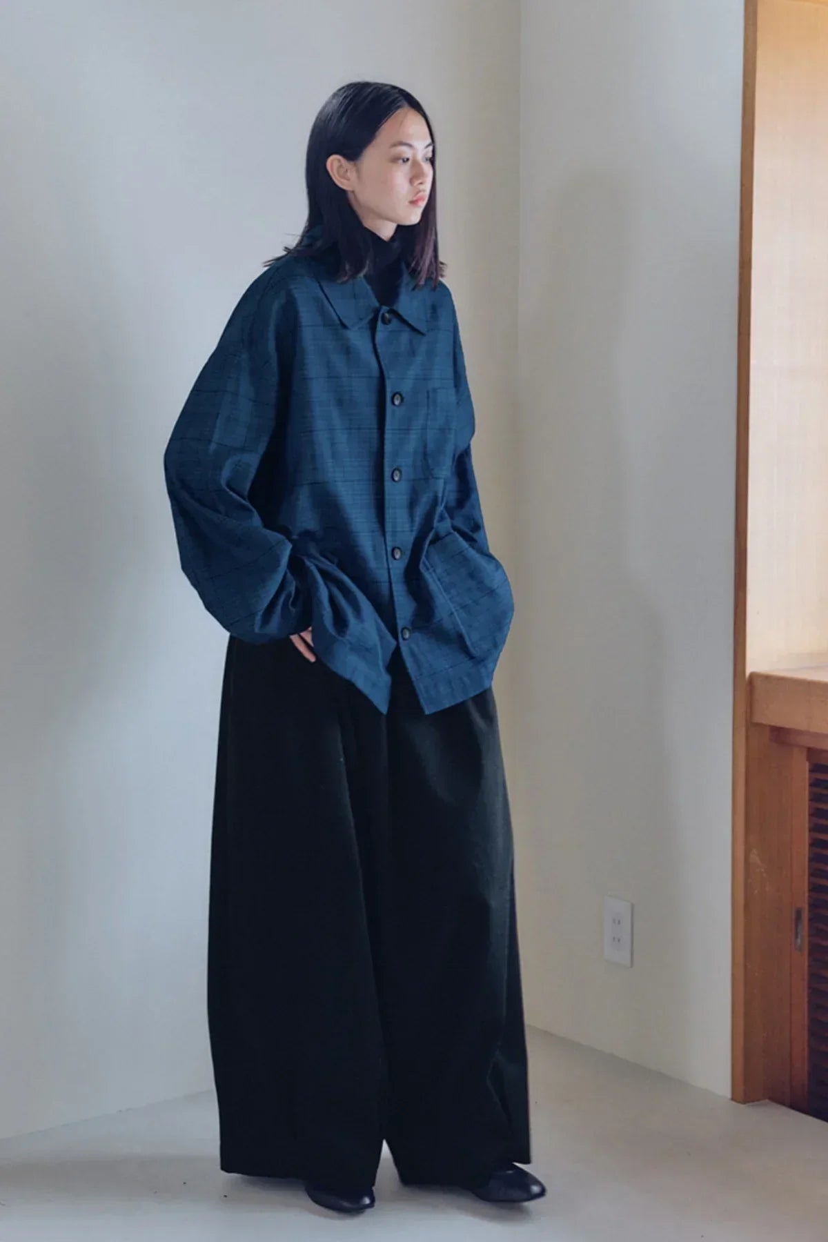 Model wearing black wide-leg unisex tuck balloon pants with a dark blue checkered oversized shirt indoors