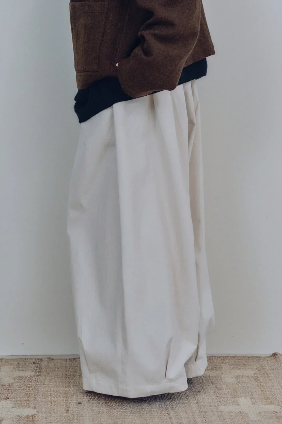 close-up of unisex cream-colored tuck balloon pants paired with brown jacket on neutral floor