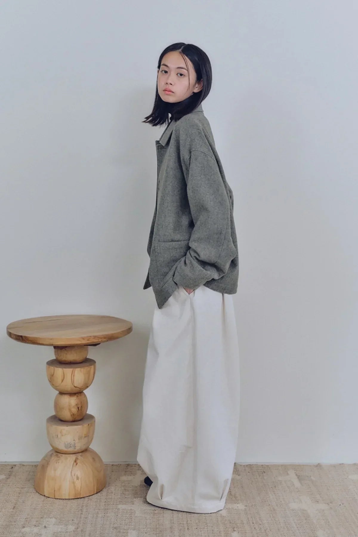 Young woman wearing oversized gray jacket and wide cream balloon pants standing next to wooden side table
