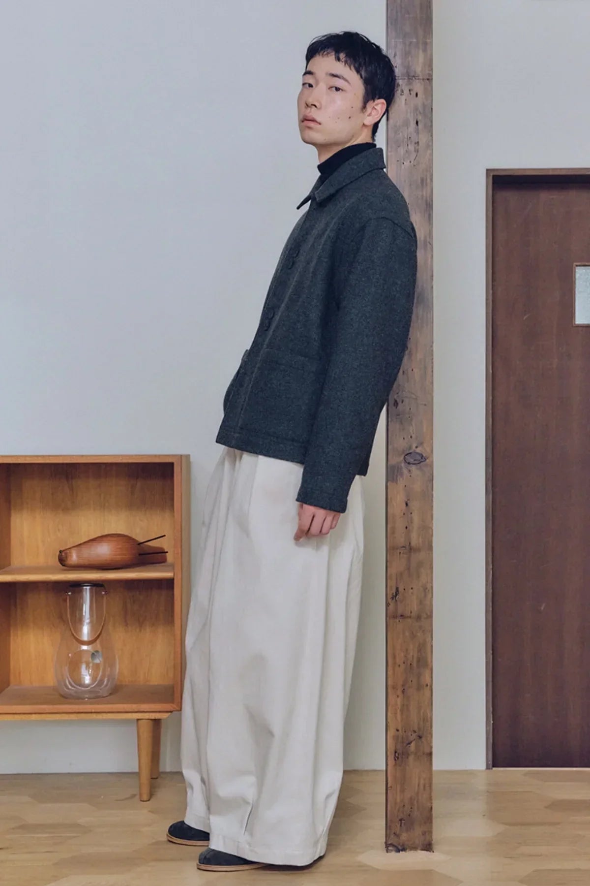 Man wearing a dark gray jacket and cream unisex tuck balloon pants standing indoors near wooden shelf and beam