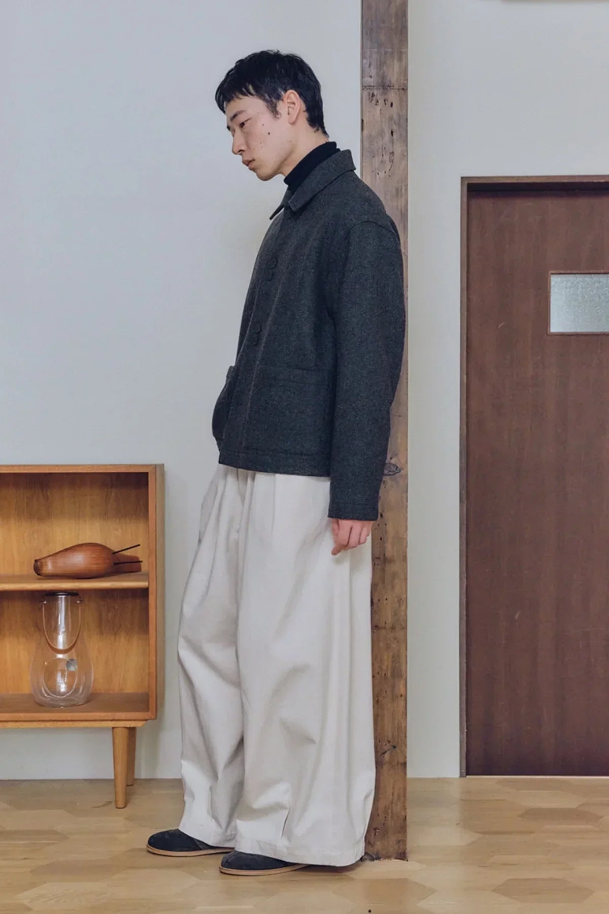 Man wearing cream wide tuck balloon pants, dark jacket, and black shoes in minimalist room
