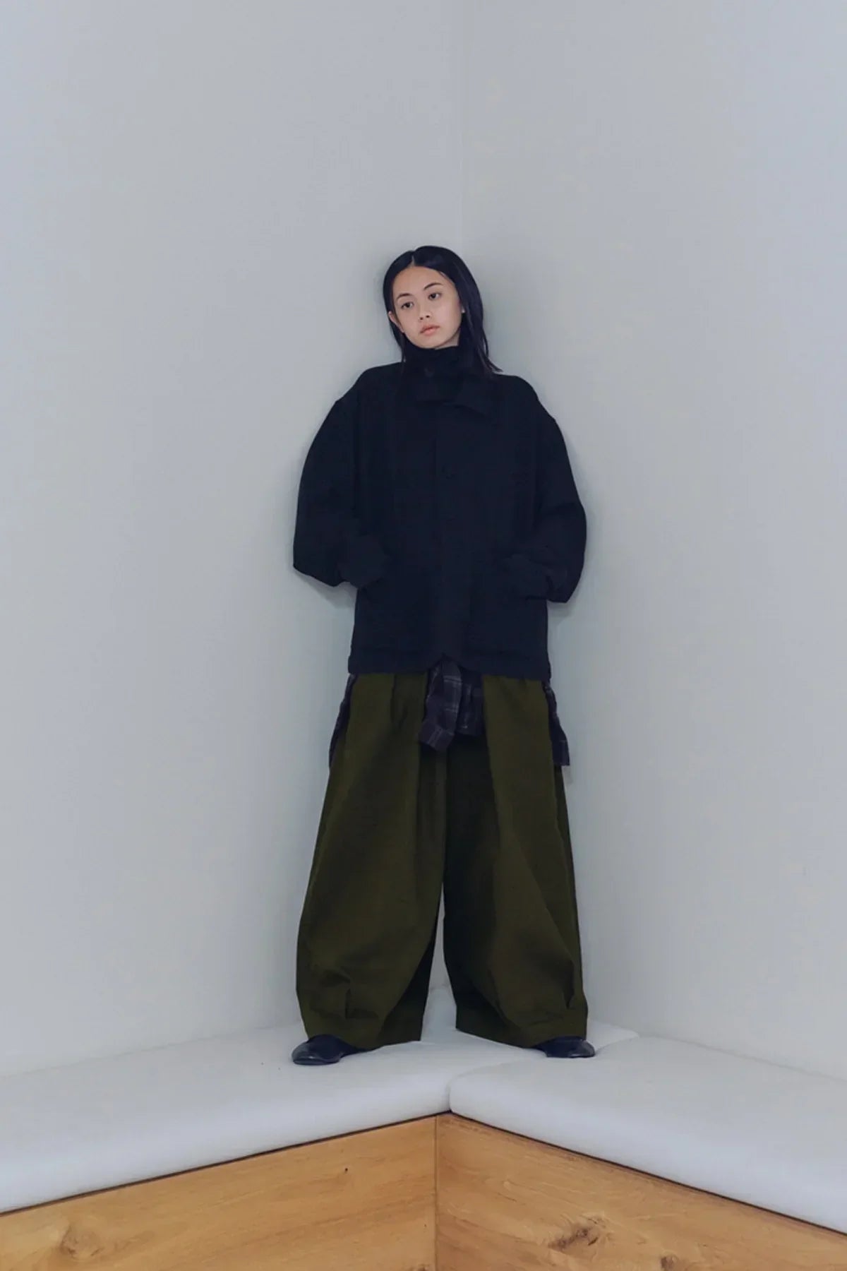 Model wearing unisex khaki balloon pants with black oversized jacket, standing in minimalist corner setting