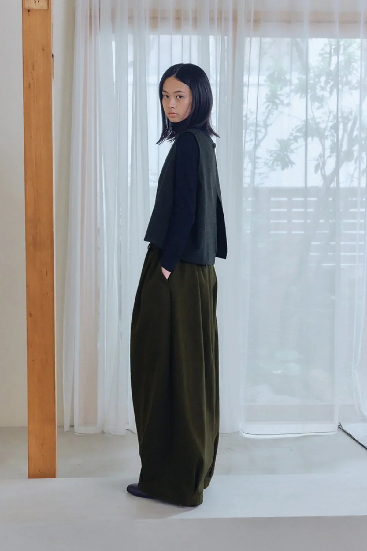 Woman wearing black top and olive green unisex tuck balloon pants in minimalist room with sheer curtains