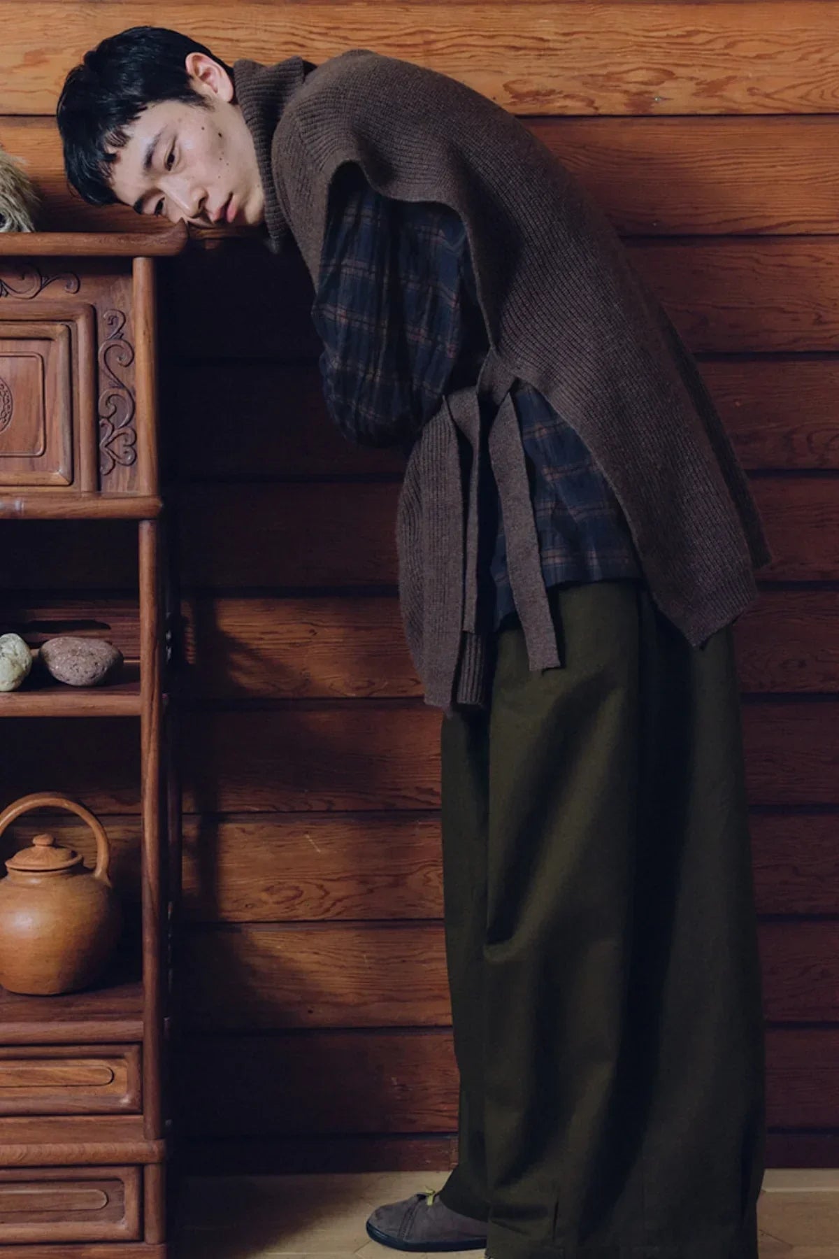 Man leaning against wooden cabinet wearing brown knit vest, plaid shirt, and olive balloon pants in rustic room