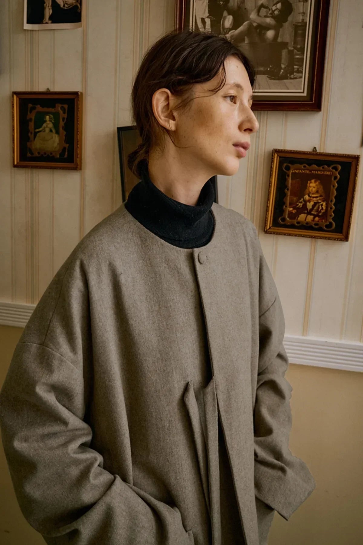 Woman wearing beige wool jacket over black turtleneck standing indoors with vintage framed art