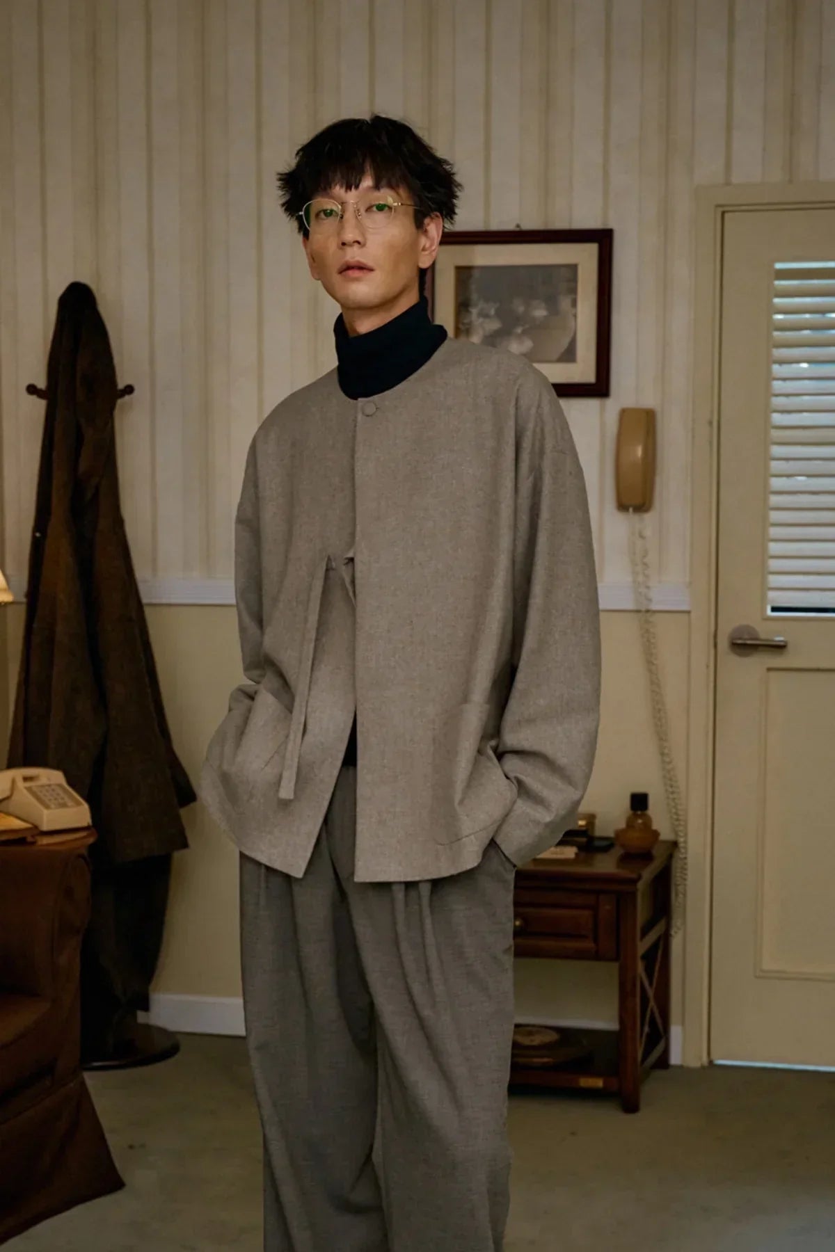 Man wearing beige wool loop jacket and gray trousers standing in vintage room with coat rack and rotary phone