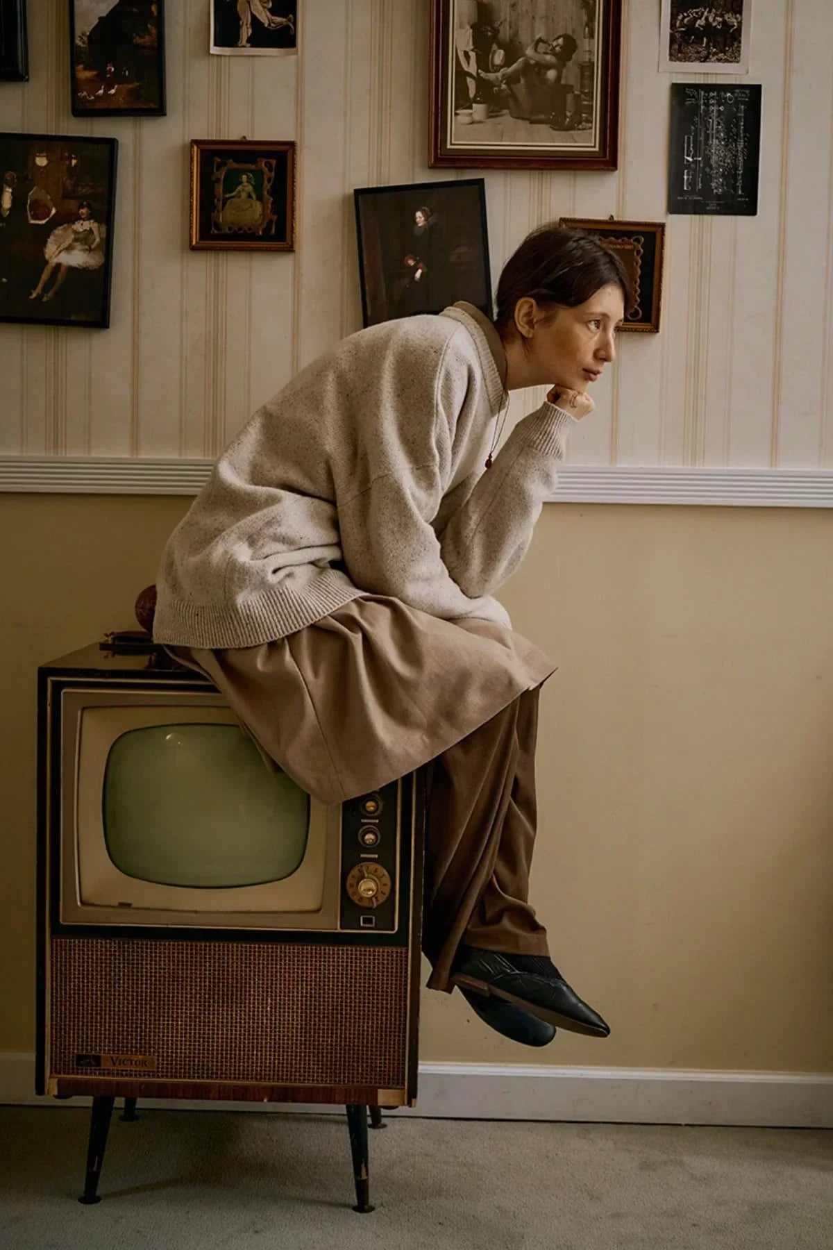 Person wearing beige wool sweater and brown pants, sitting on vintage Victor TV in retro room