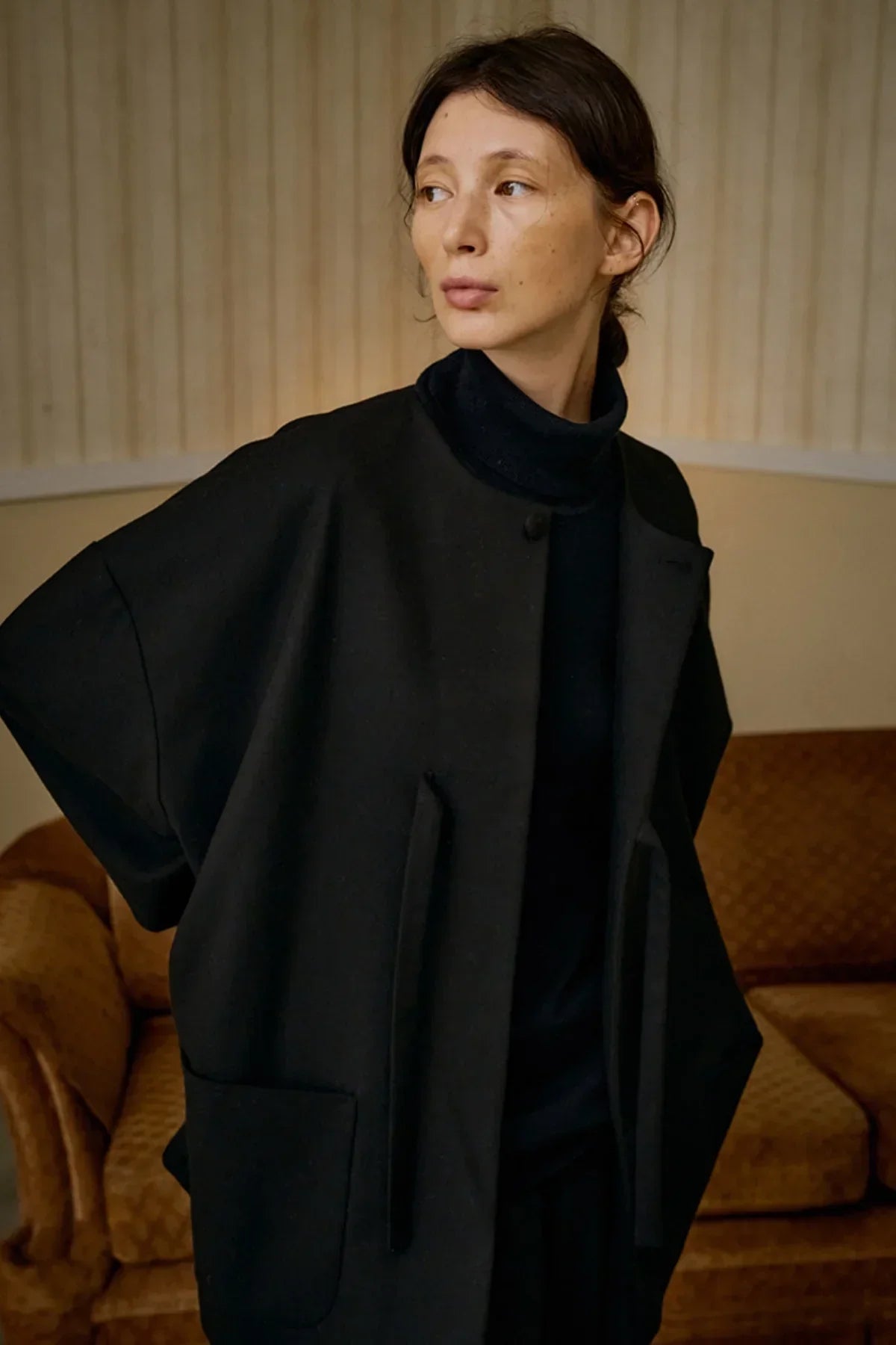 Woman wearing black wool loop jacket over black turtleneck indoors with brown sofa background