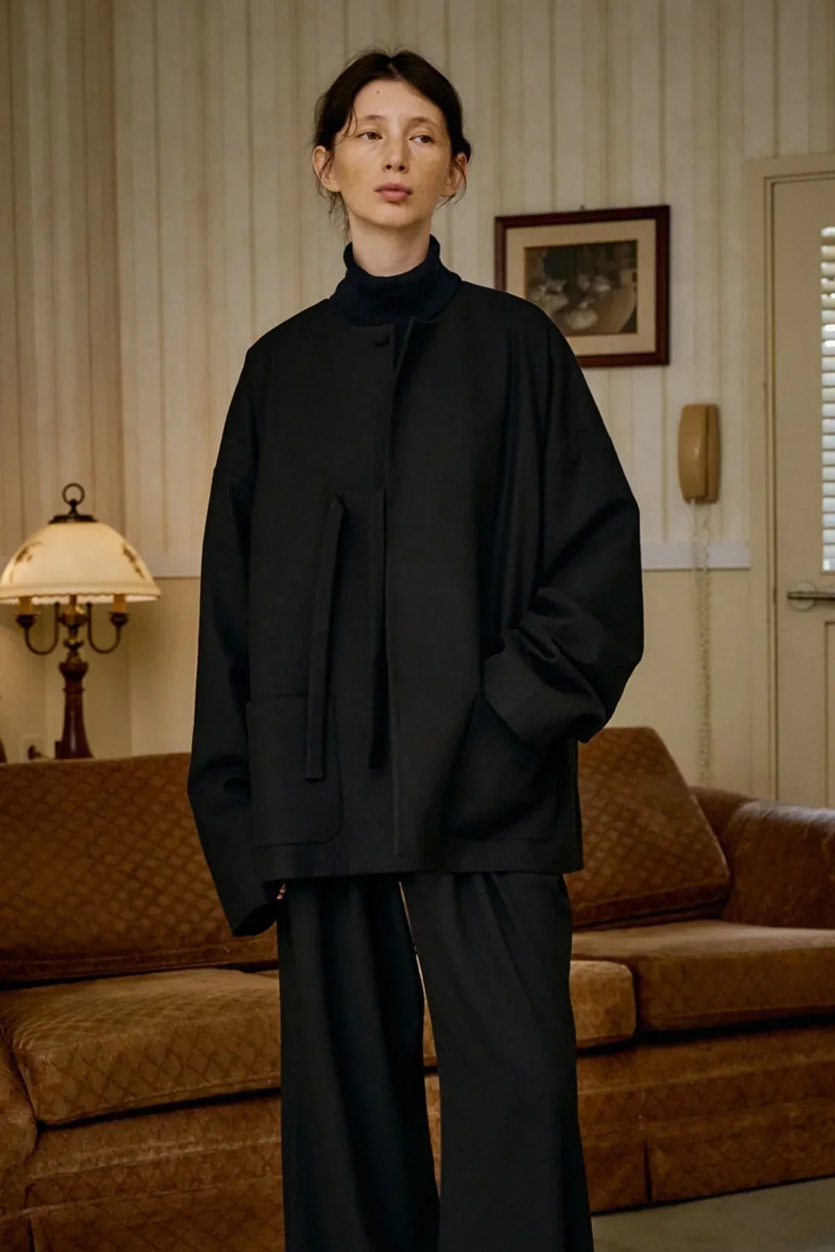 Model wearing black unisex wool loop jacket with pockets, standing in vintage-style living room