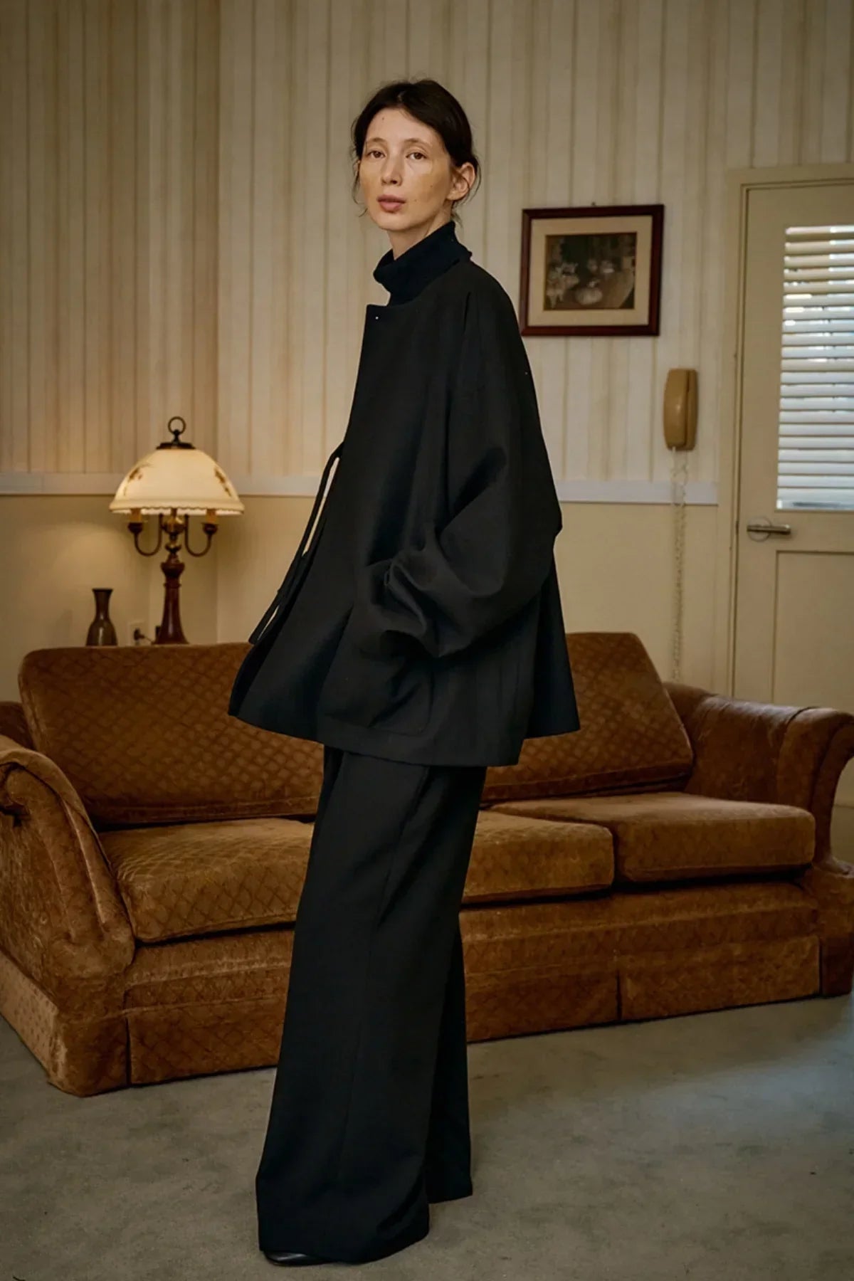 Model wearing black unisex wool loop jacket and wide-leg pants in vintage-style living room