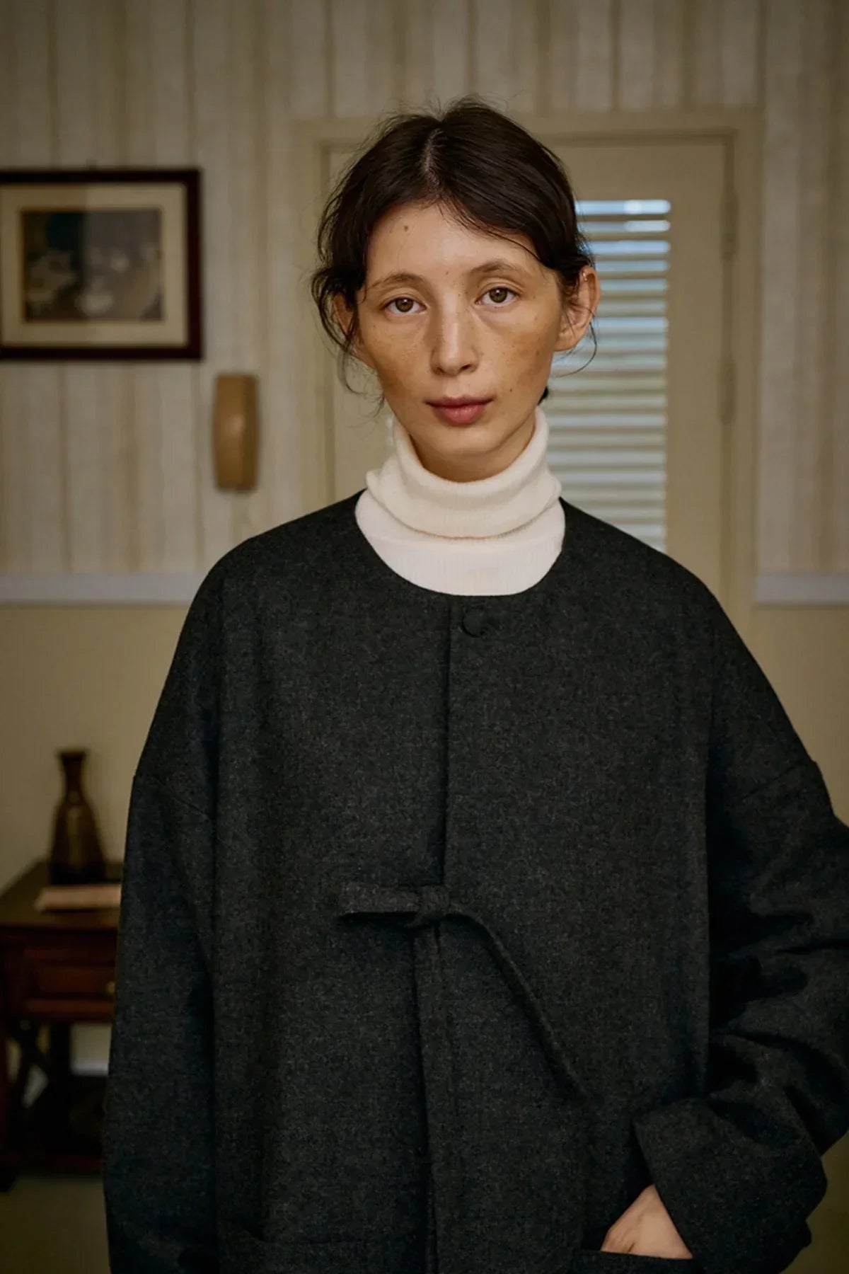Close-up of a person wearing a charcoal unisex wool loop jacket over a white turtleneck indoors