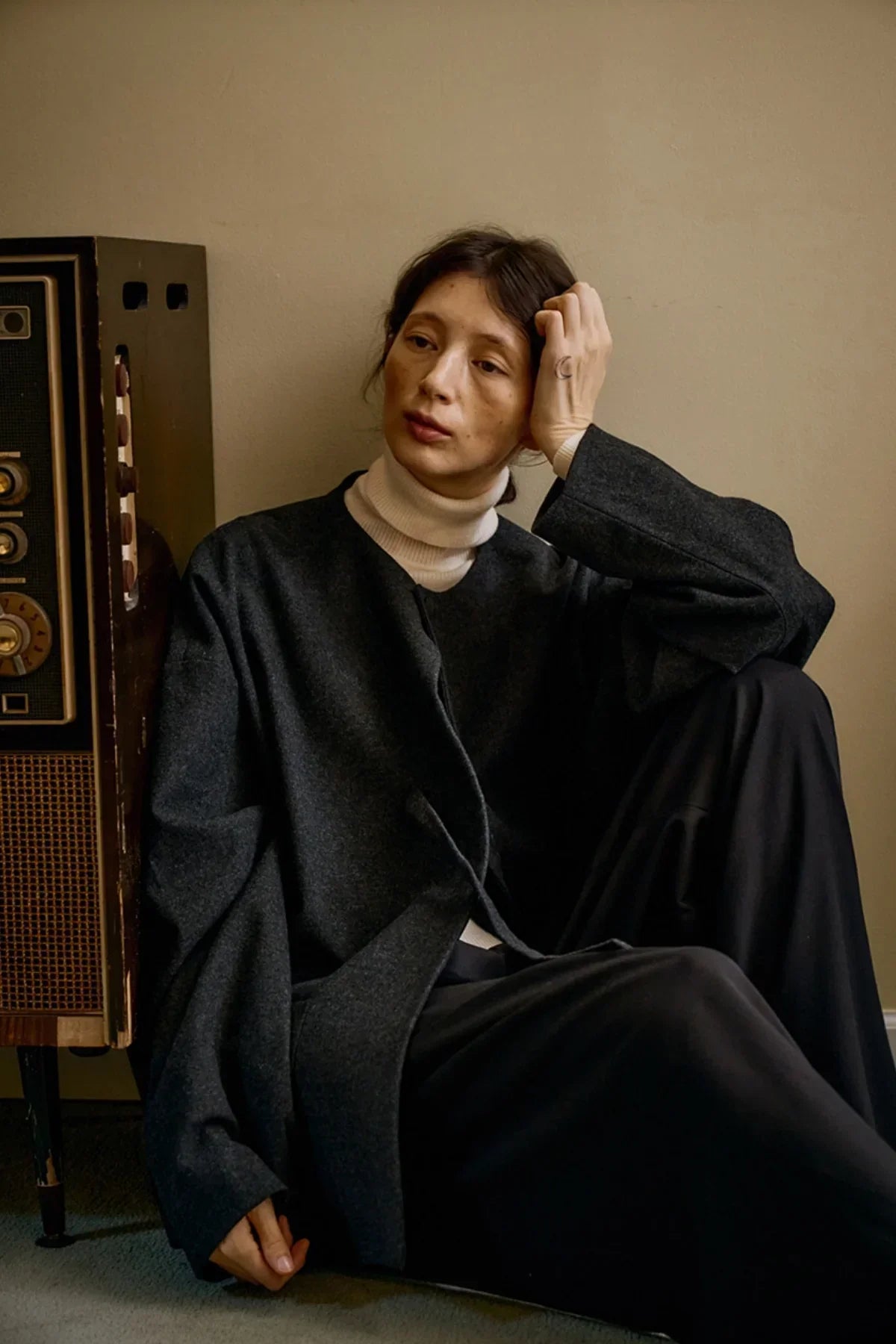 Person wearing a charcoal unisex wool loop jacket and white turtleneck, sitting beside a vintage radio