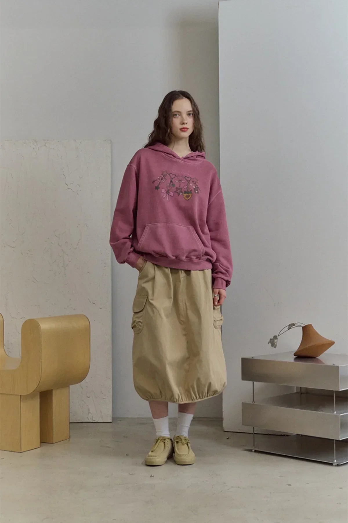 Woman wearing dusty pink graphic hoodie and beige balloon skirt in minimalist room with wooden chair and modern decor