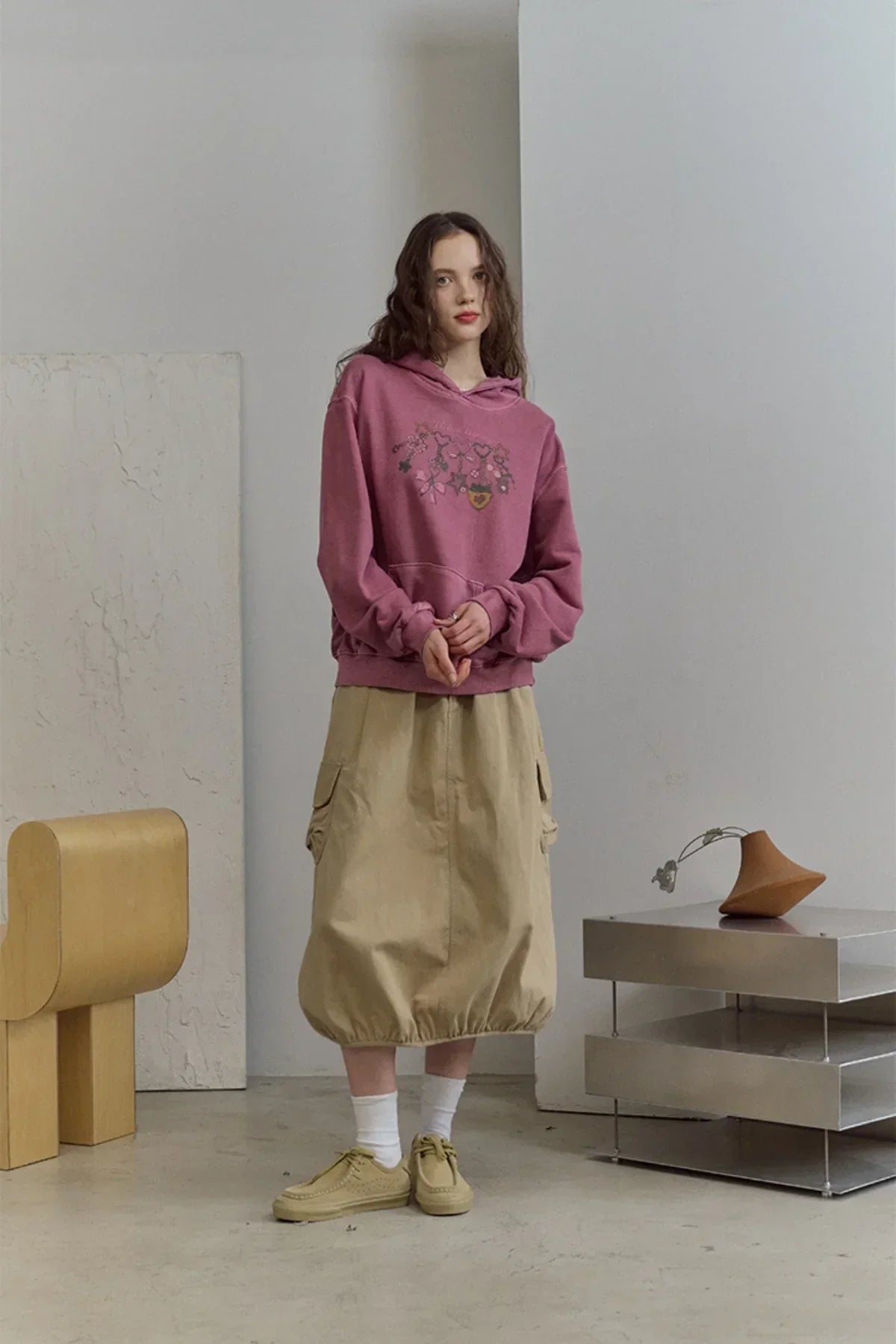 Young woman in dusty pink floral hoodie, beige cargo skirt, white socks, and beige shoes in minimalist room