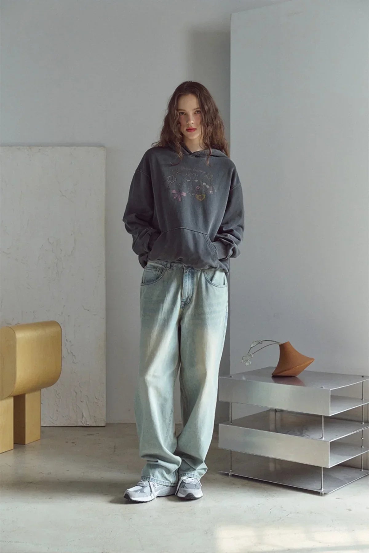 Model wearing smoke black pigment hoodie with graphic print, light washed jeans, and sneakers in minimalist room