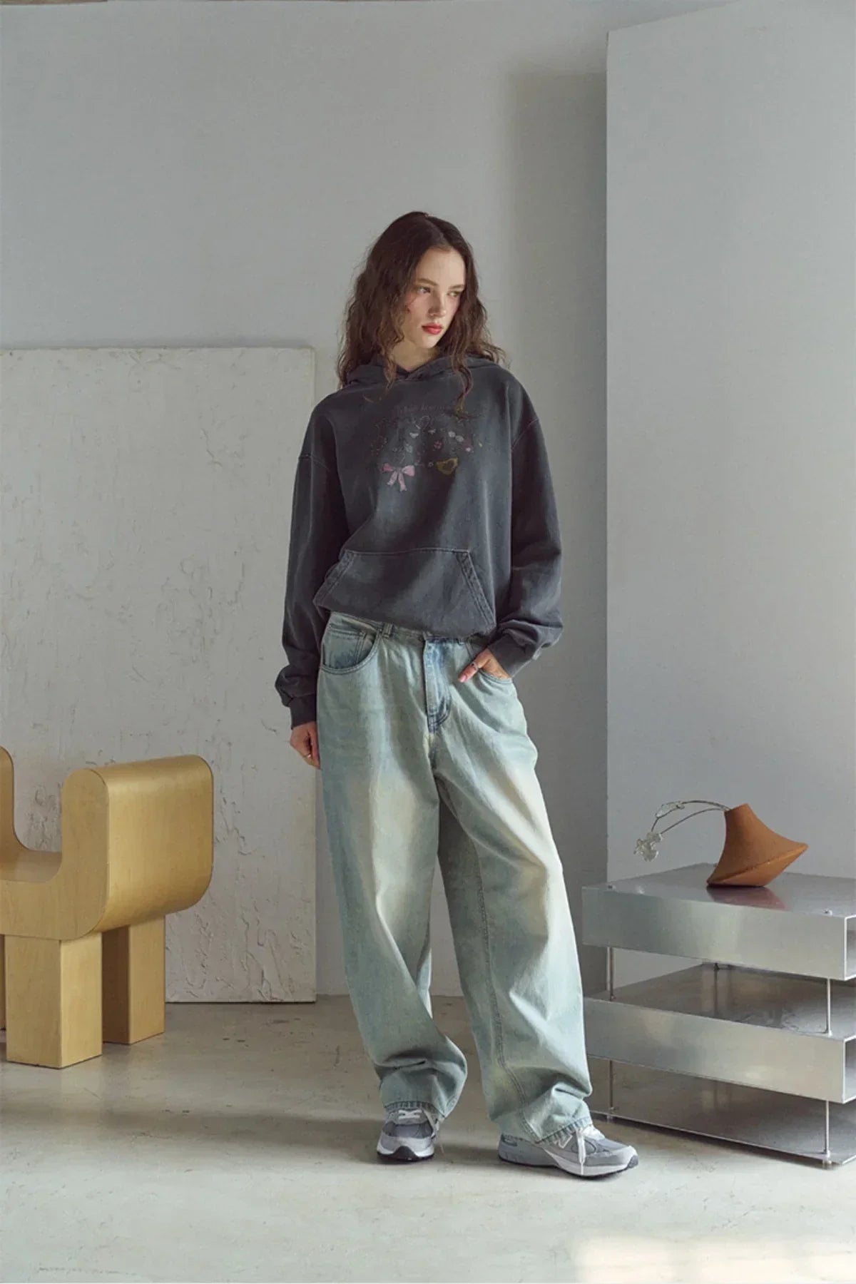 Woman in loose light-wash jeans, dark graphic hoodie, and sneakers standing in minimalist room