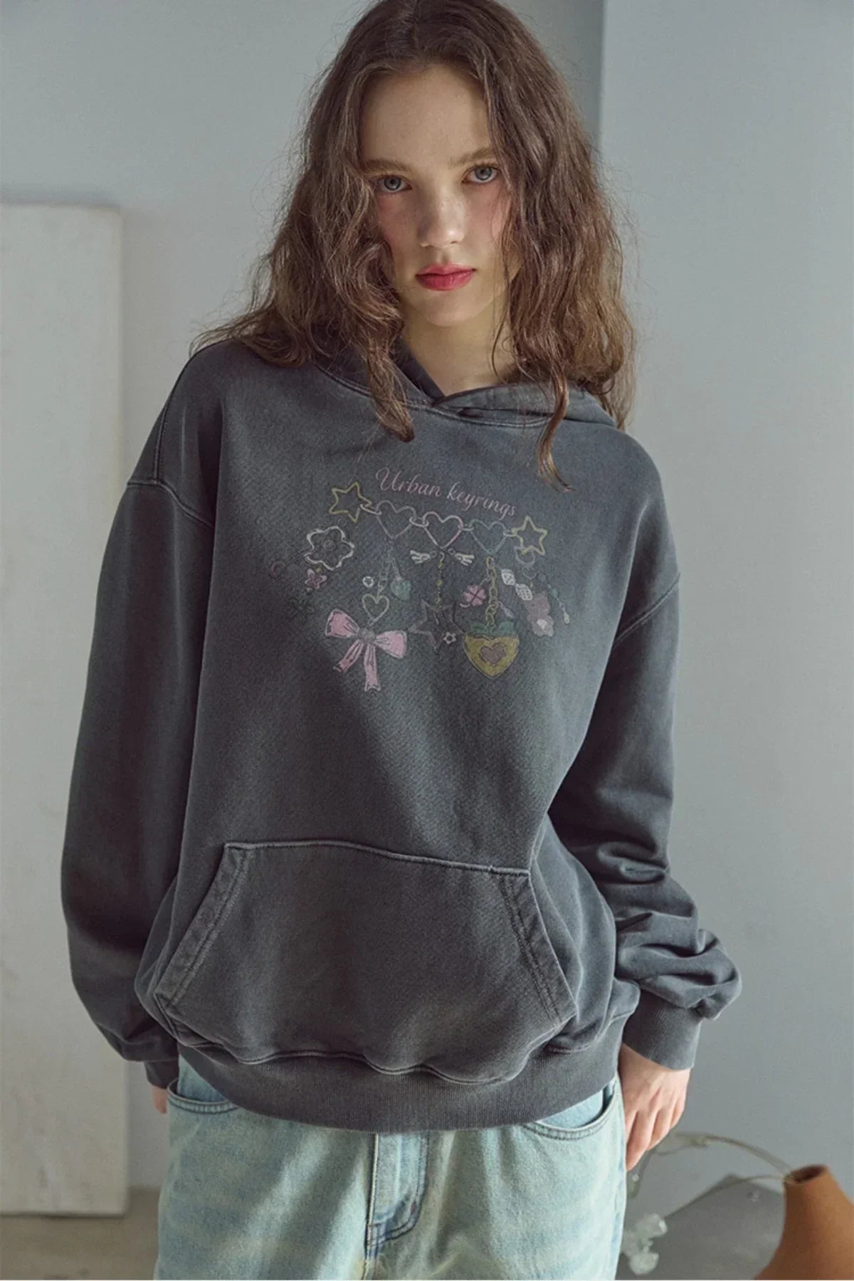 Young woman wearing a smoke black hoodie with colorful keyring graphic and light blue jeans in minimal indoor setting
